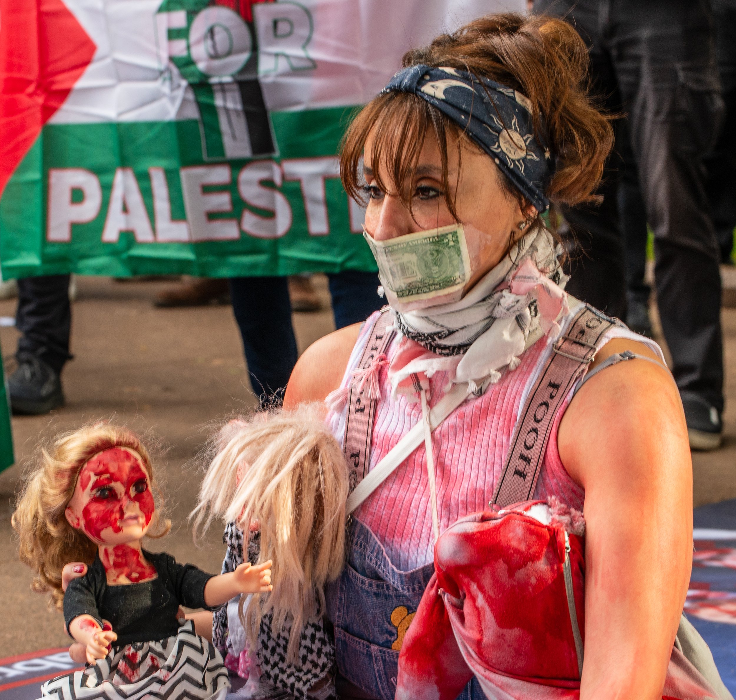 A woman with a bandana on her head, a scarf covering her mouth with a dollar bill printed on it, and carrying a doll with blood on her face and a stuffed animal. In the background, a Palestinian flag is visible that reads 'FOR PALESTINE'.