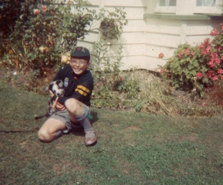 Young boy wearing glasses, a black and yellow cap, black jacket, shorts, and purple shoes, kneeling on the grass holding a small dog with a leash in front of a house with bushes and flowers.