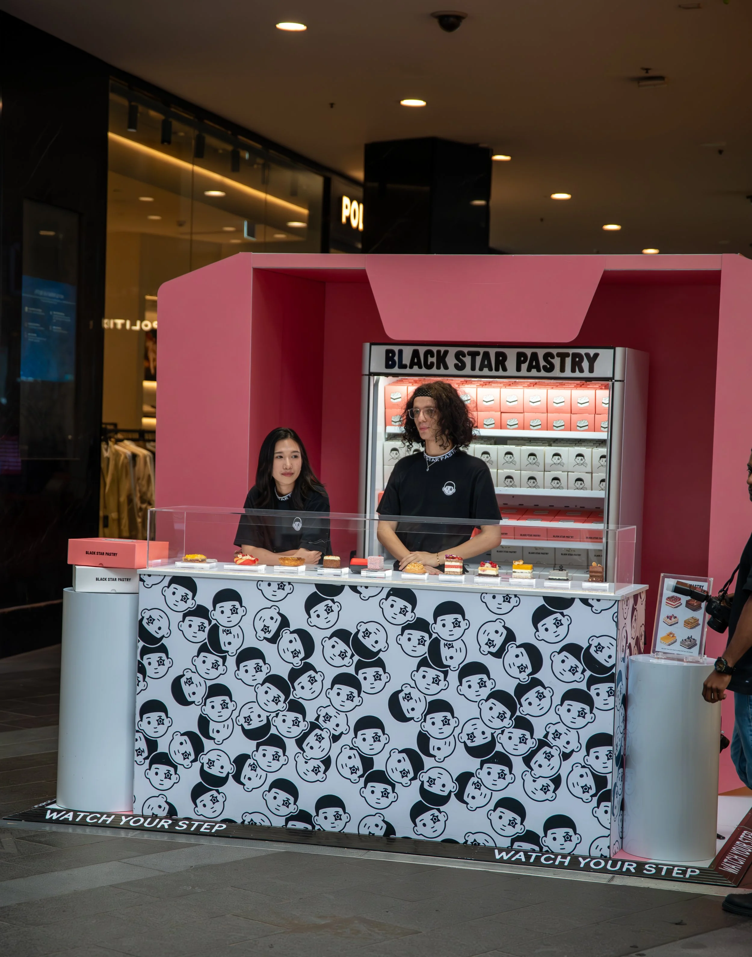 A bakery kiosk named Black Star Pastry selling various slices of cakes in a shopping mall. The kiosk features a pink and black design with cartoon faces on the front panel and a glass display case showcasing the cakes.