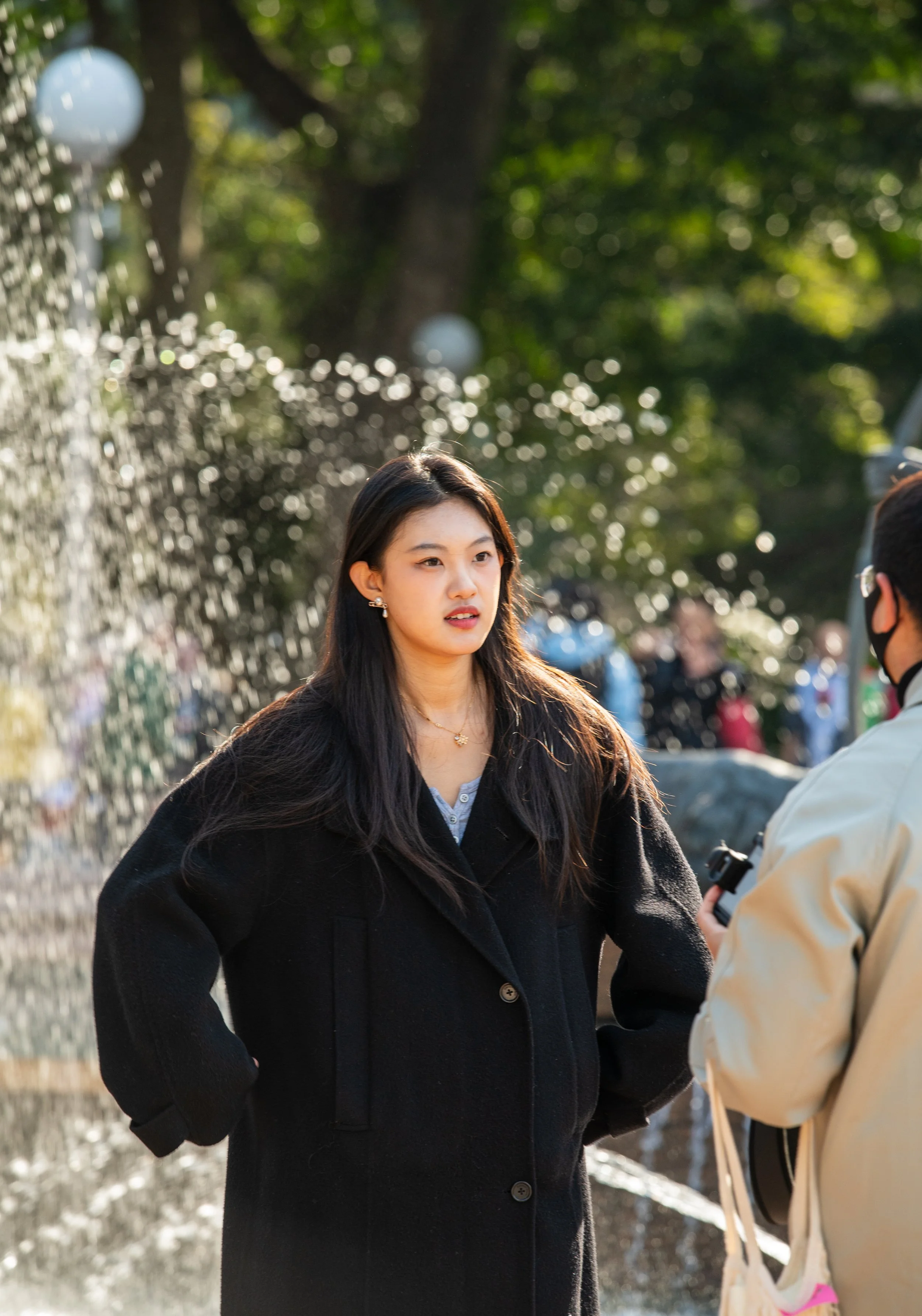 A young woman with long dark hair, wearing a black coat, stands outdoors near a water fountain with sprays of water, while a person next to her, partly visible, holds a camera. There are blurred people and greenery in the background.