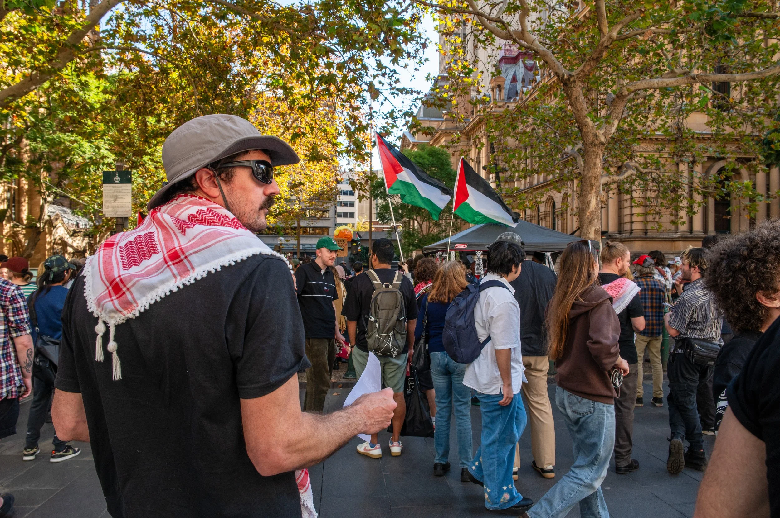 A man with sunglasses and a hat, wearing a scarf and holding a paper, stands at a gathering with a crowd and flags in the background.