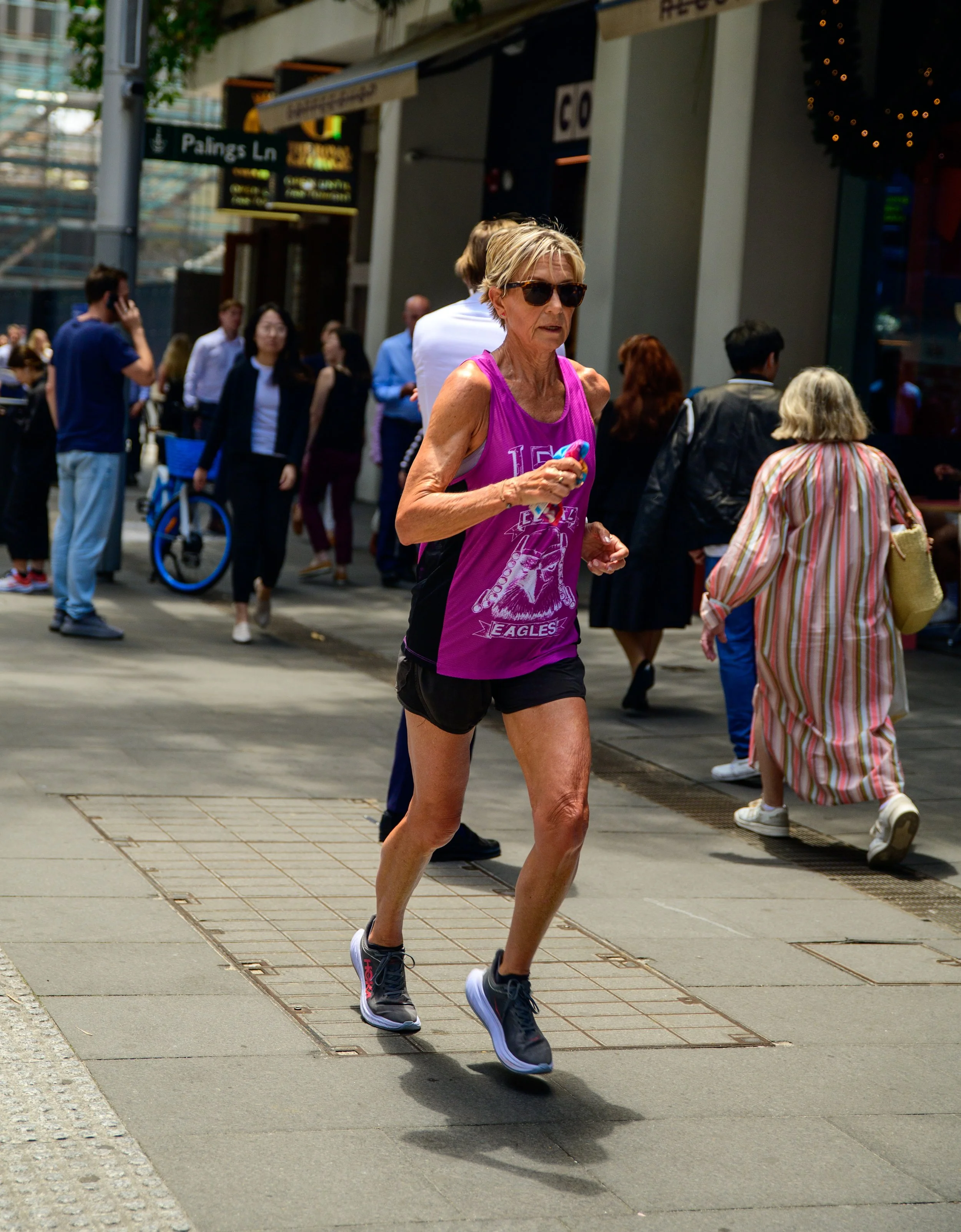 An older woman running on a city sidewalk during a public event or race, wearing sunglasses, a purple athletic tank top, black shorts, and running shoes.