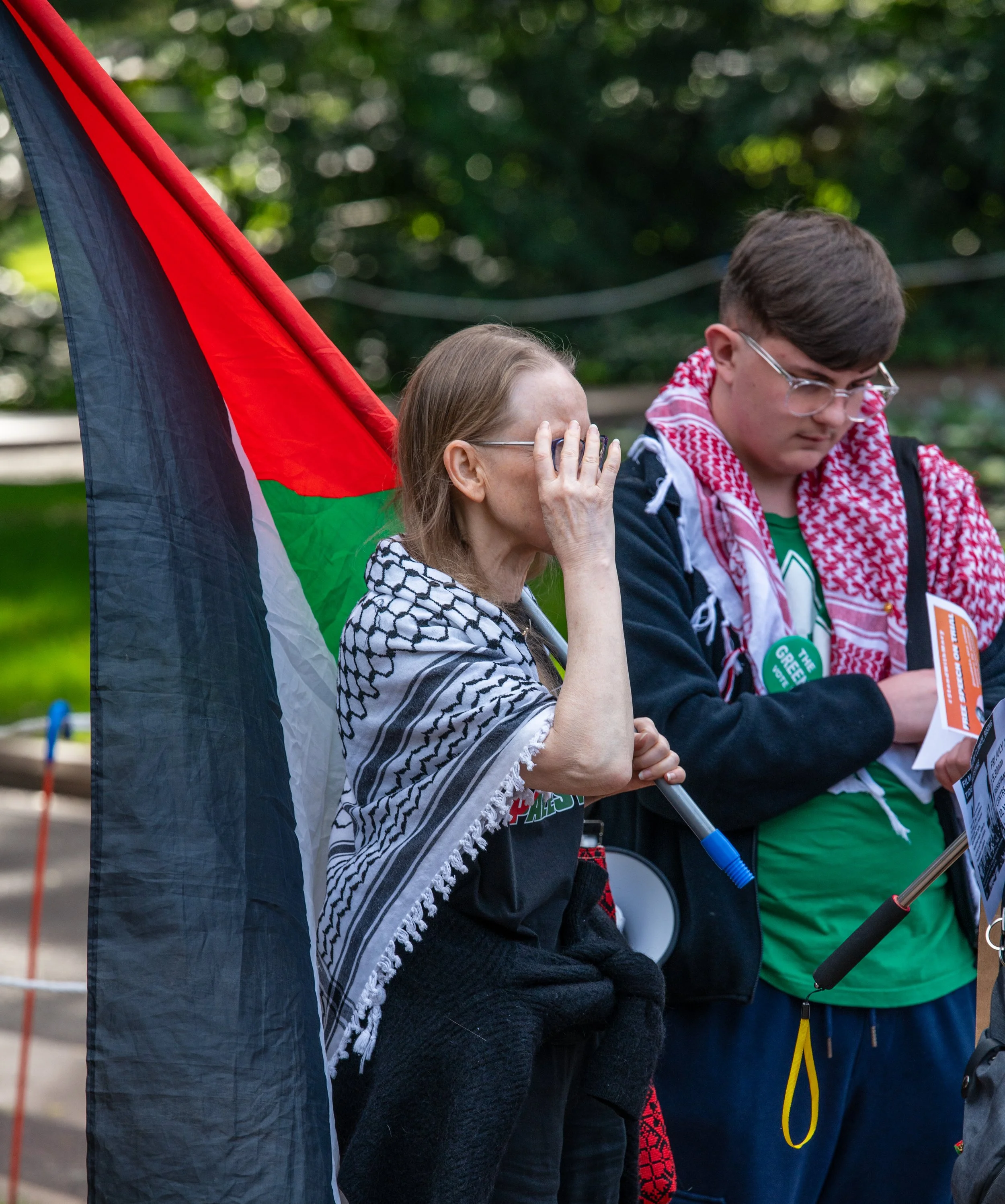 A woman with a turnout scarf on her shoulders, standing next to a young man in glasses, both involved in a protest or demonstration. The woman has her hand on her face, and there is a Palestinian flag behind them.
