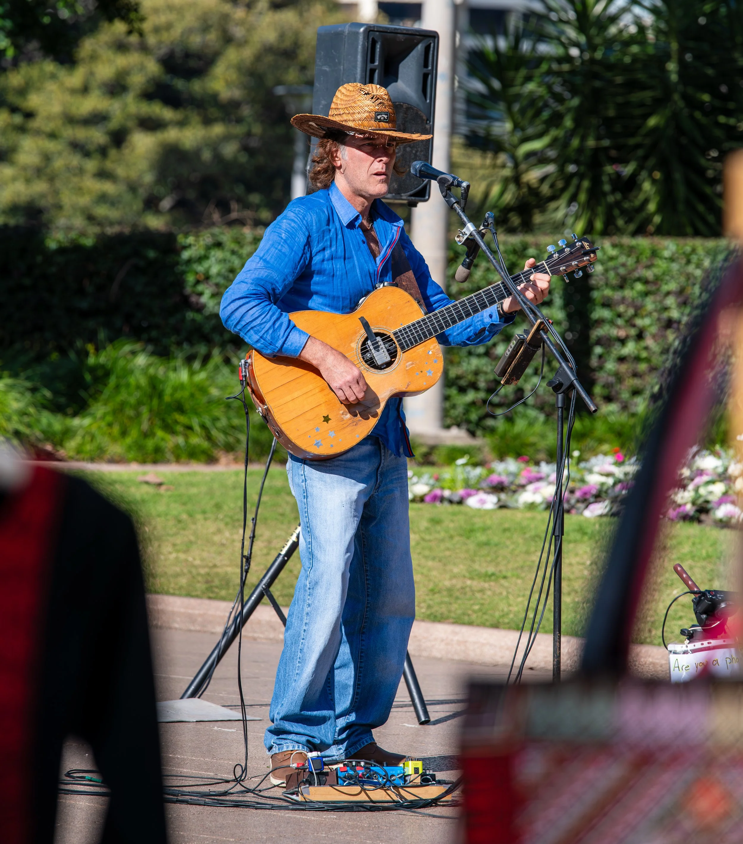 A man wearing a straw hat and a blue shirt playing an acoustic guitar during an outdoor performance, standing in front of a microphone and sound equipment with greenery and flowers in the background.