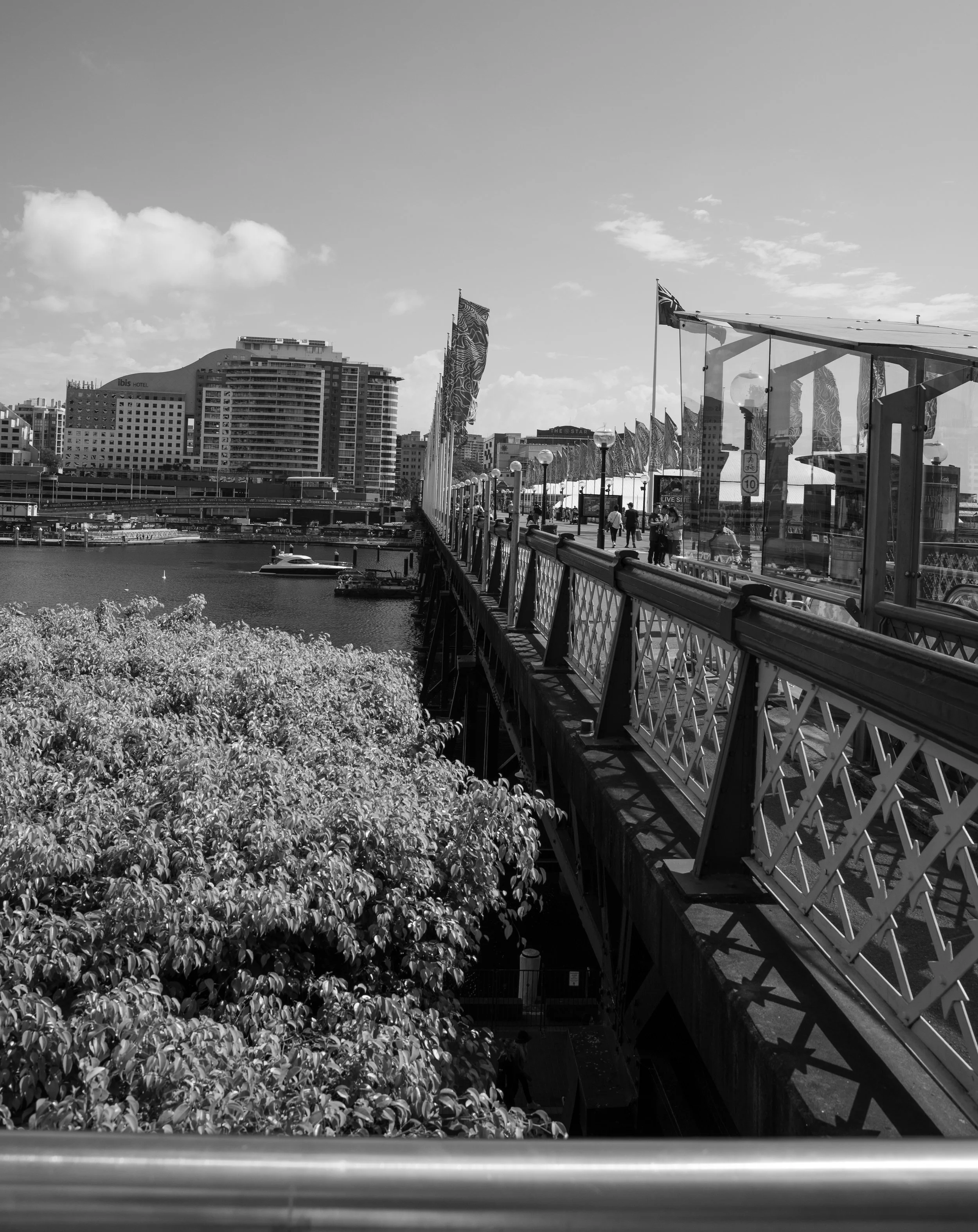 A black and white photo of a pedestrian bridge over a river in an urban area, with buildings, flags, and people walking.