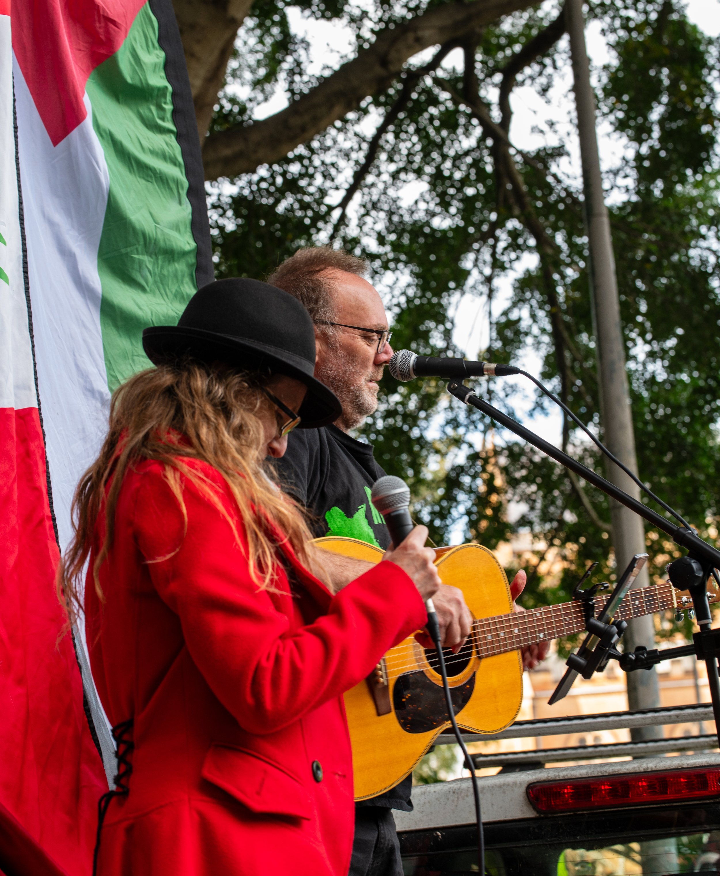 A woman in a red coat and black hat singing into a microphone and a man playing guitar and singing into a microphone, standing outdoors near a flag with red, white, green, and black colors.