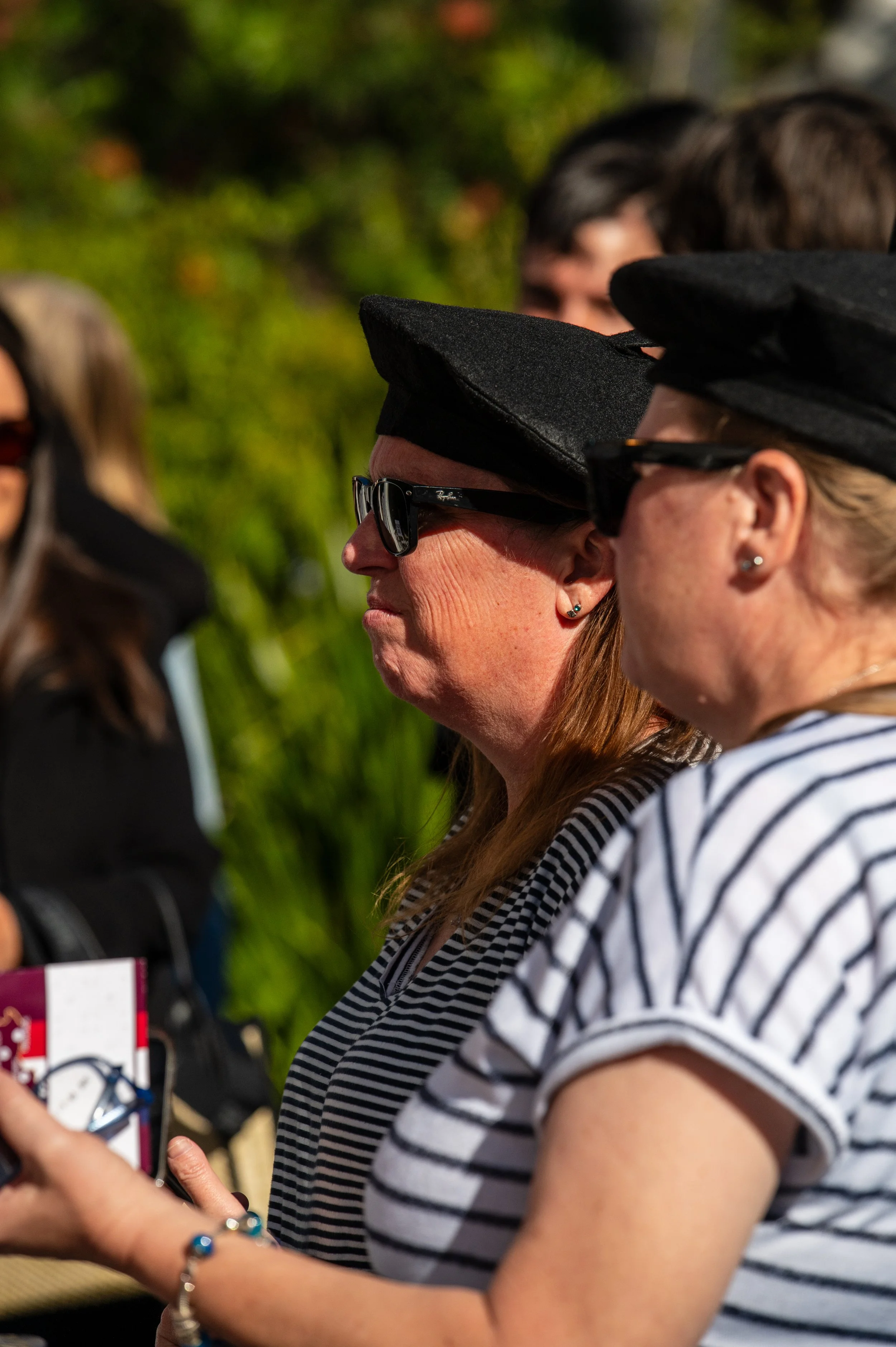 Two women wearing black hats and sunglasses standing outdoors, one in a striped shirt holding a phone.