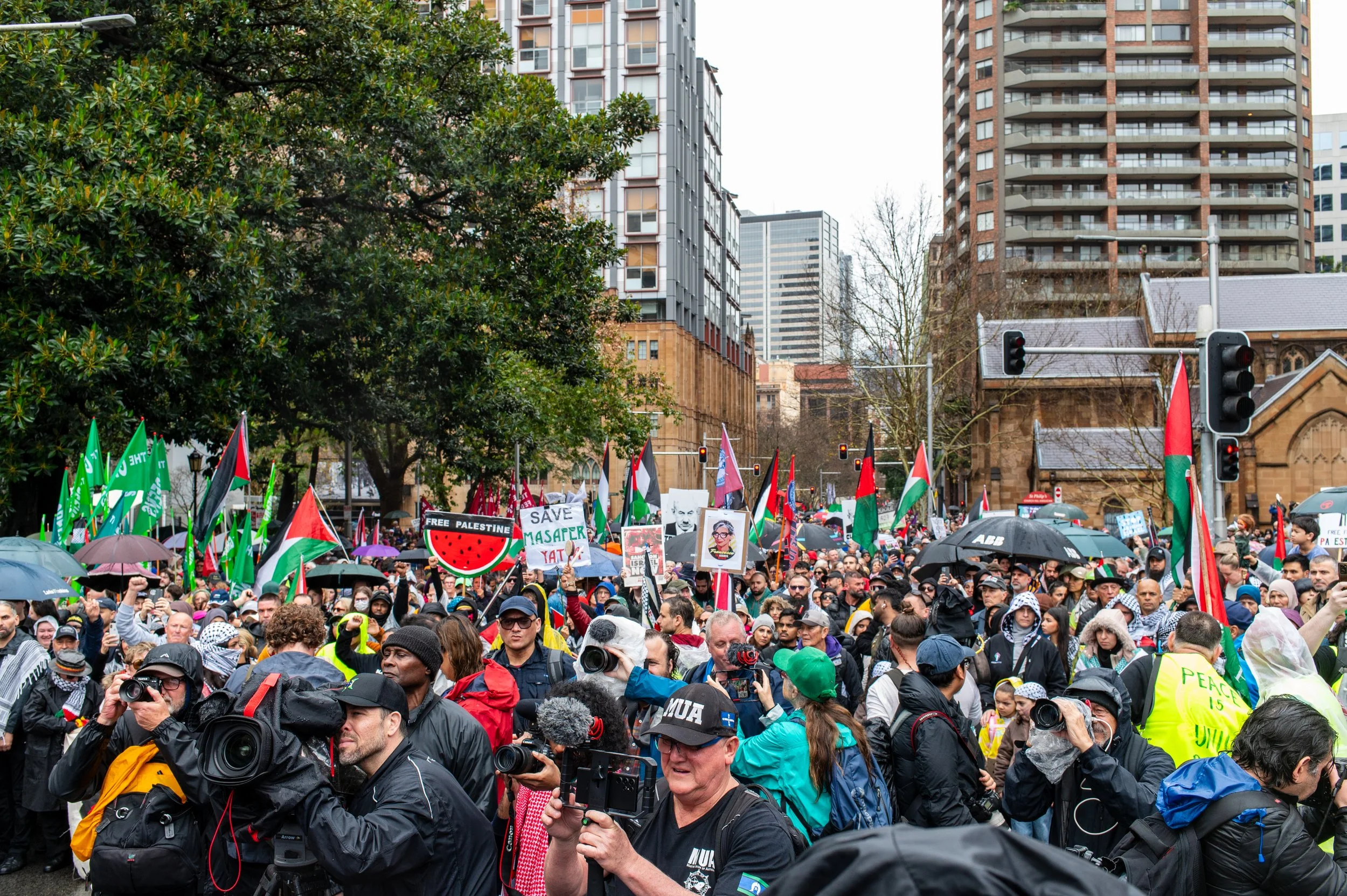 A large crowd of protesters holding flags, signs, and umbrellas during a demonstration in an urban area with tall buildings and trees.