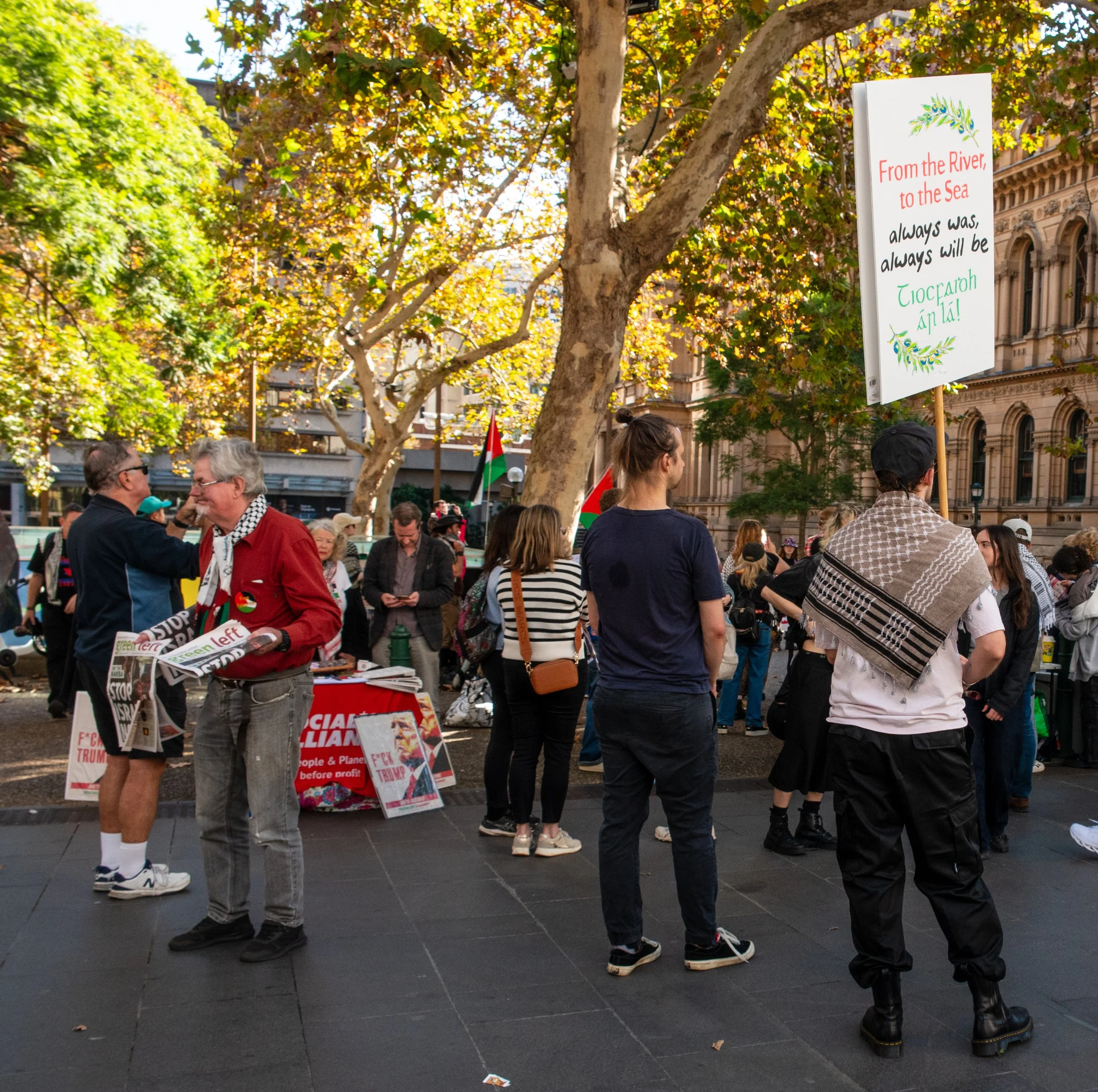 People gathered outdoors in a park or city square, some holding signs and banners, with trees and historic buildings in the background, during a protest or demonstration.