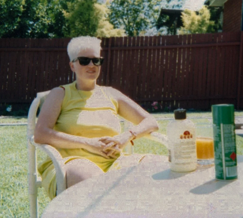 Person with short white hair wearing sunglasses and a yellow dress sitting outside at a table with condiments and drinks.