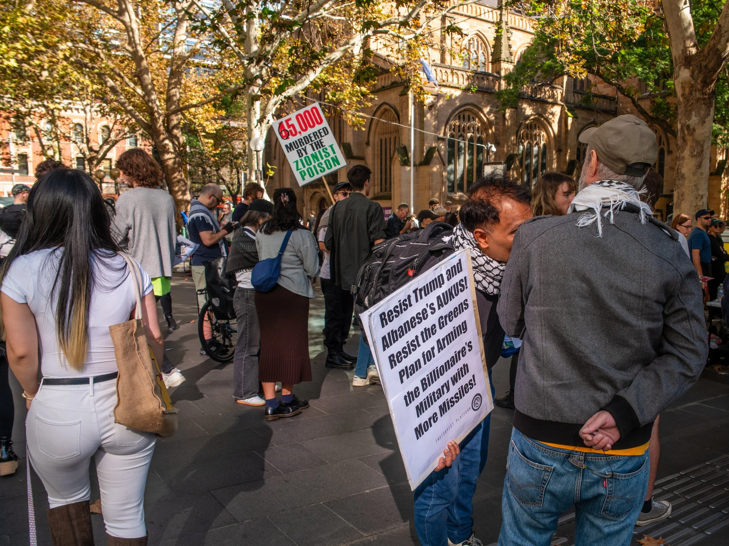 A group of protesters at an outdoor rally holding signs that oppose political violence and express resistance against certain political figures, with trees and historic buildings in the background.