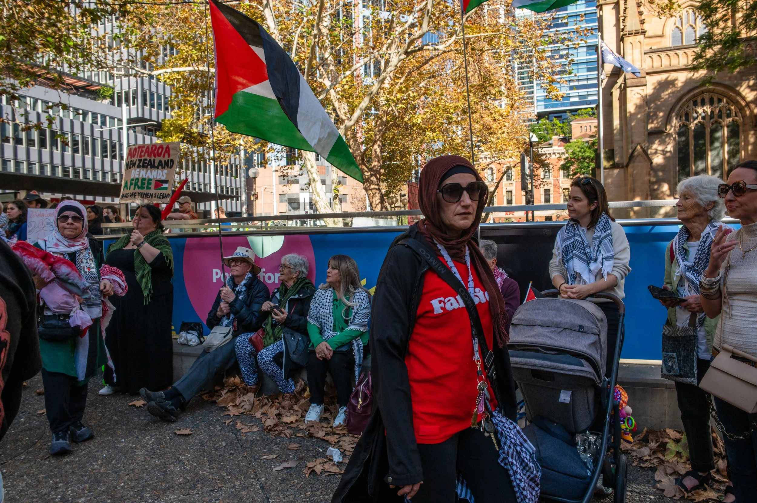 Group of women gathered outdoors during a protest or rally, some holding a Palestinian flag and a sign demanding free New Zealand. The scene is set in an urban area with trees and buildings in the background.
