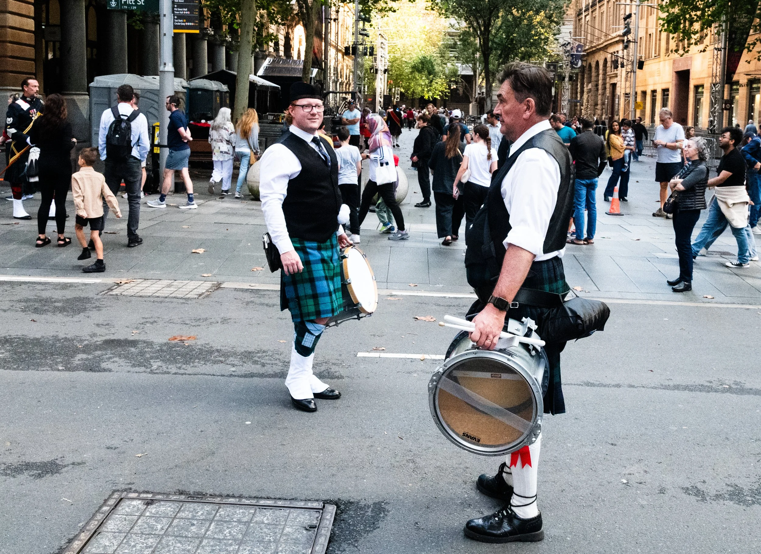 Two men dressed in traditional Scottish attire with kilts and sporrans, holding drums, standing in the middle of a busy urban street with people walking by and city buildings in the background.