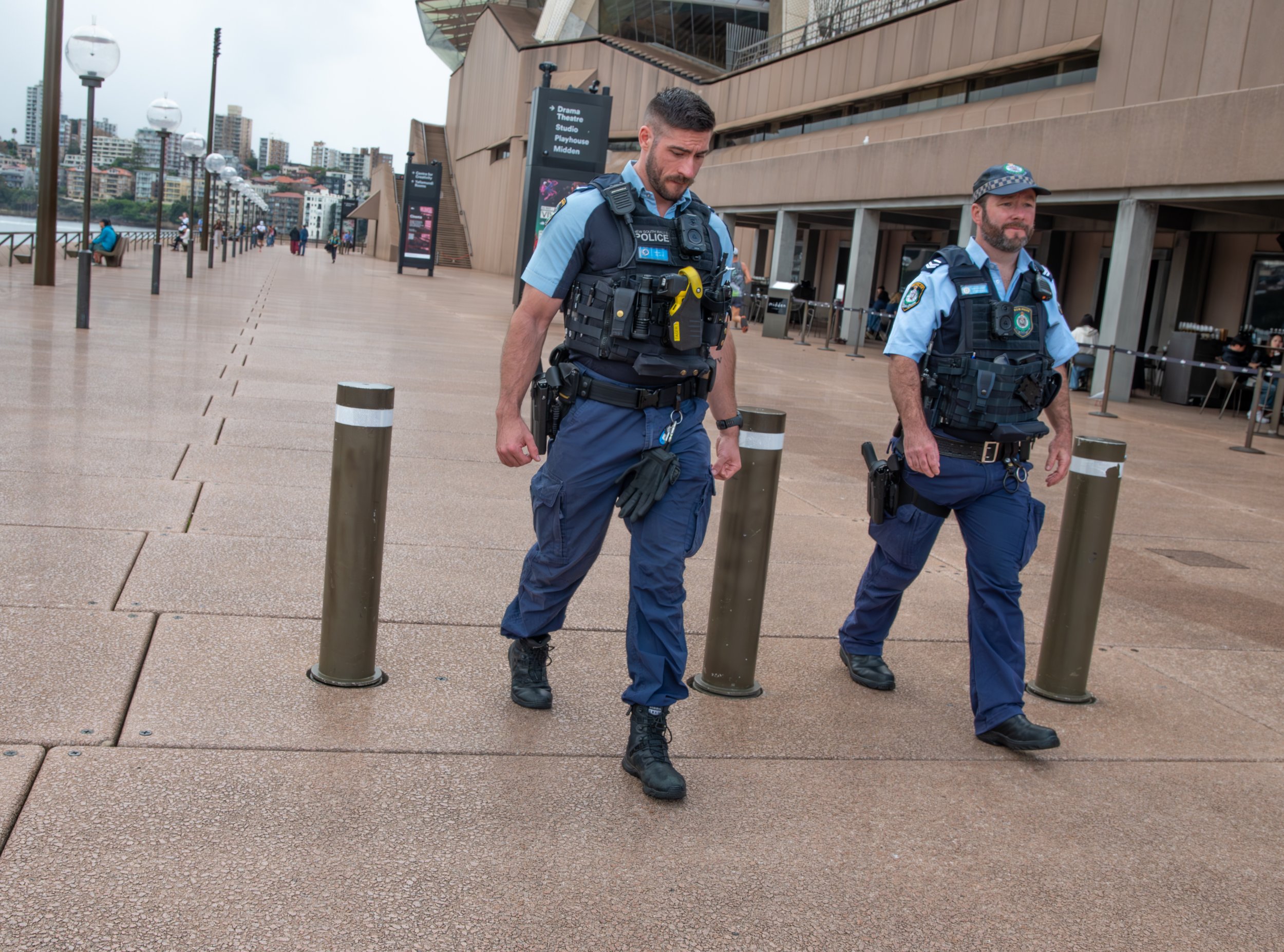 Two police officers patrol in an urban area with tall buildings and a waterfront, following a cordoned-off section with metal posts and a sidewalk.