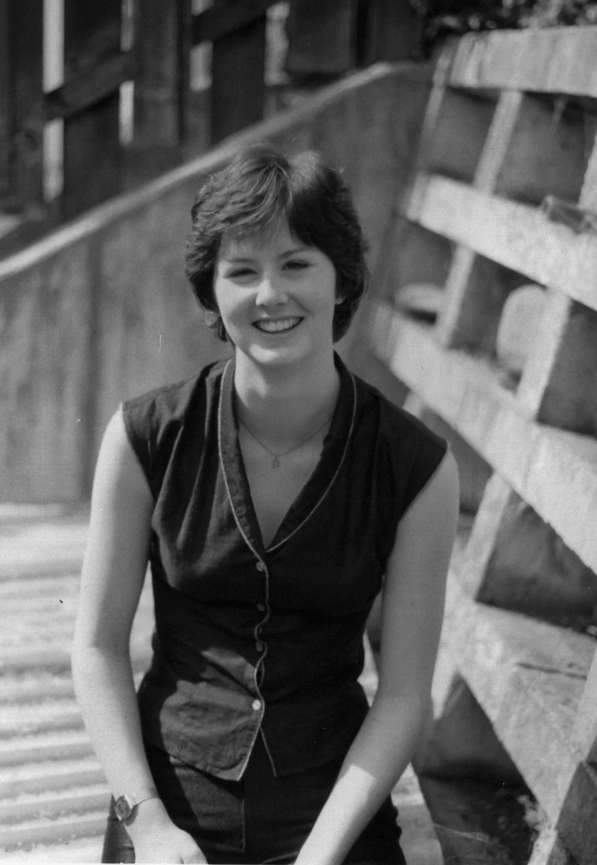 Young woman with short hair smiling, sitting outdoors near a wooden structure, wearing a sleeveless top and headphones.