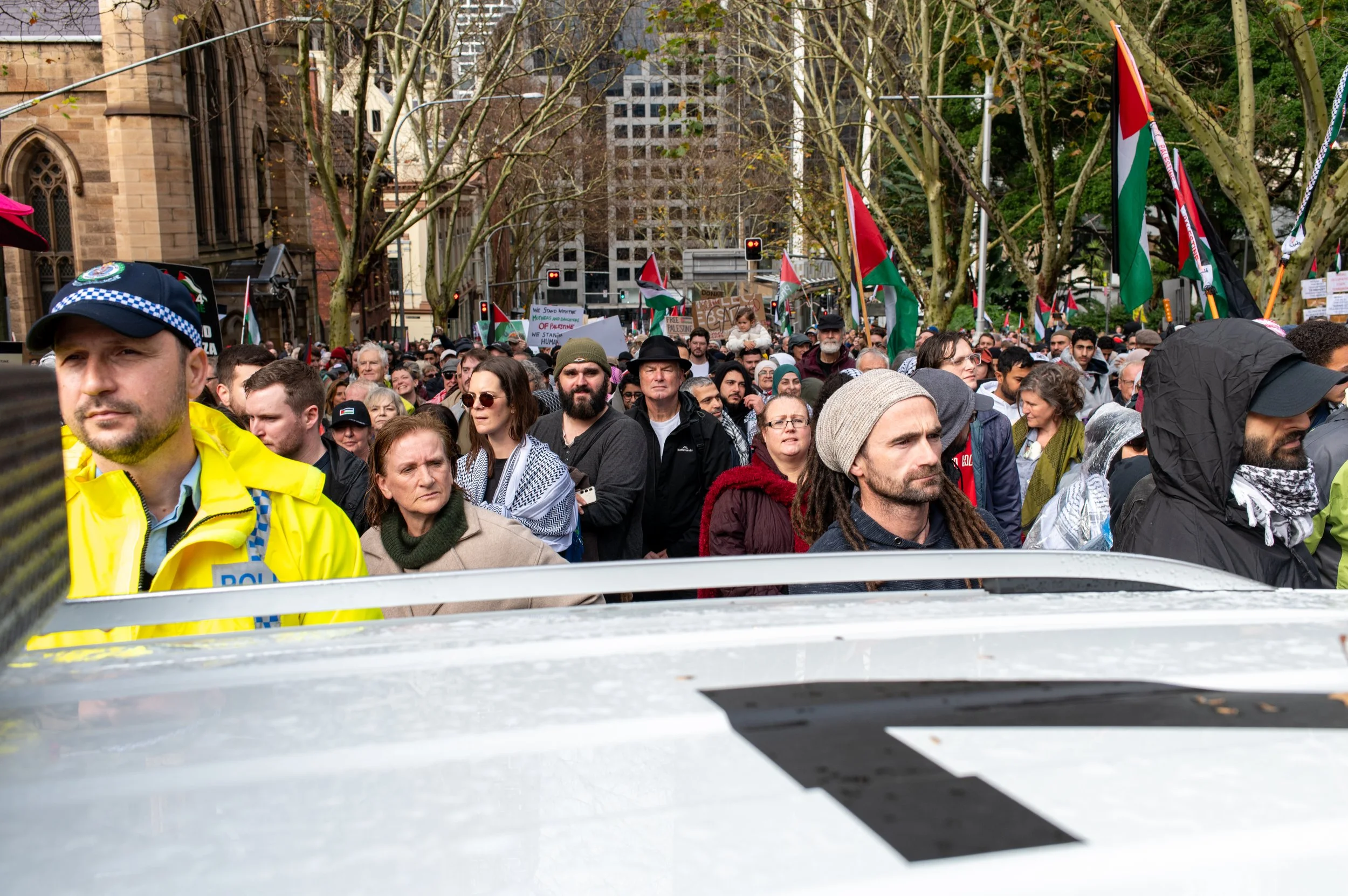 Crowd of people participating in a protest or rally on a city street, with many holding Palestinian flags. A police officer in a yellow jacket and cap is visible in the foreground, and trees line the street with buildings in the background.
