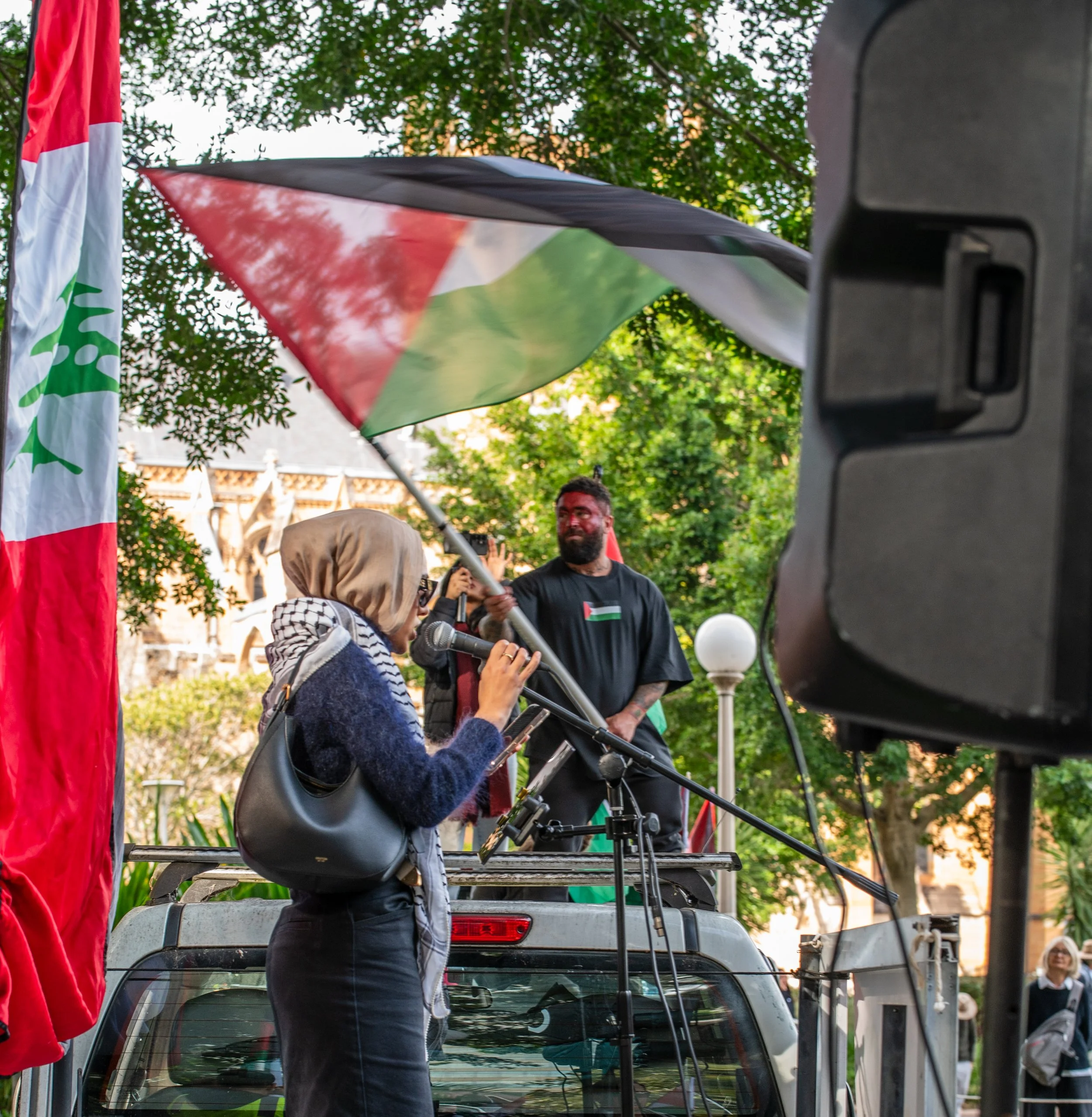 A woman with a hijab speaking into a microphone at a rally or protest, with a man standing on a vehicle behind her. The scene includes flags, one with green, black, white, and red stripes, and a person holding a large umbrella with the same colors. The setting is outdoors surrounded by trees and people.
