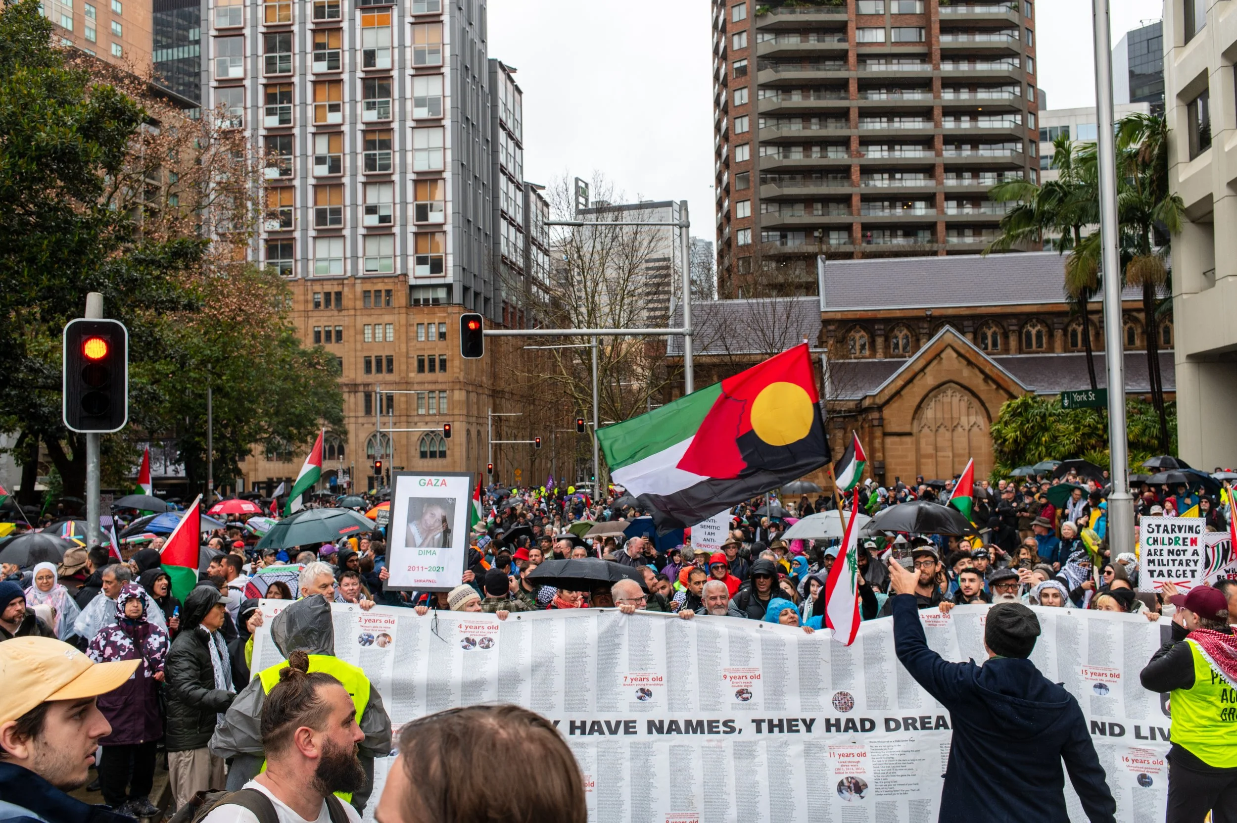 A large crowd of people gather in a city street holding signs and flags, with a prominent Palestinian flag visible. Many are using umbrellas, and some are wearing headscarves or hats. The background features tall buildings and a small church.