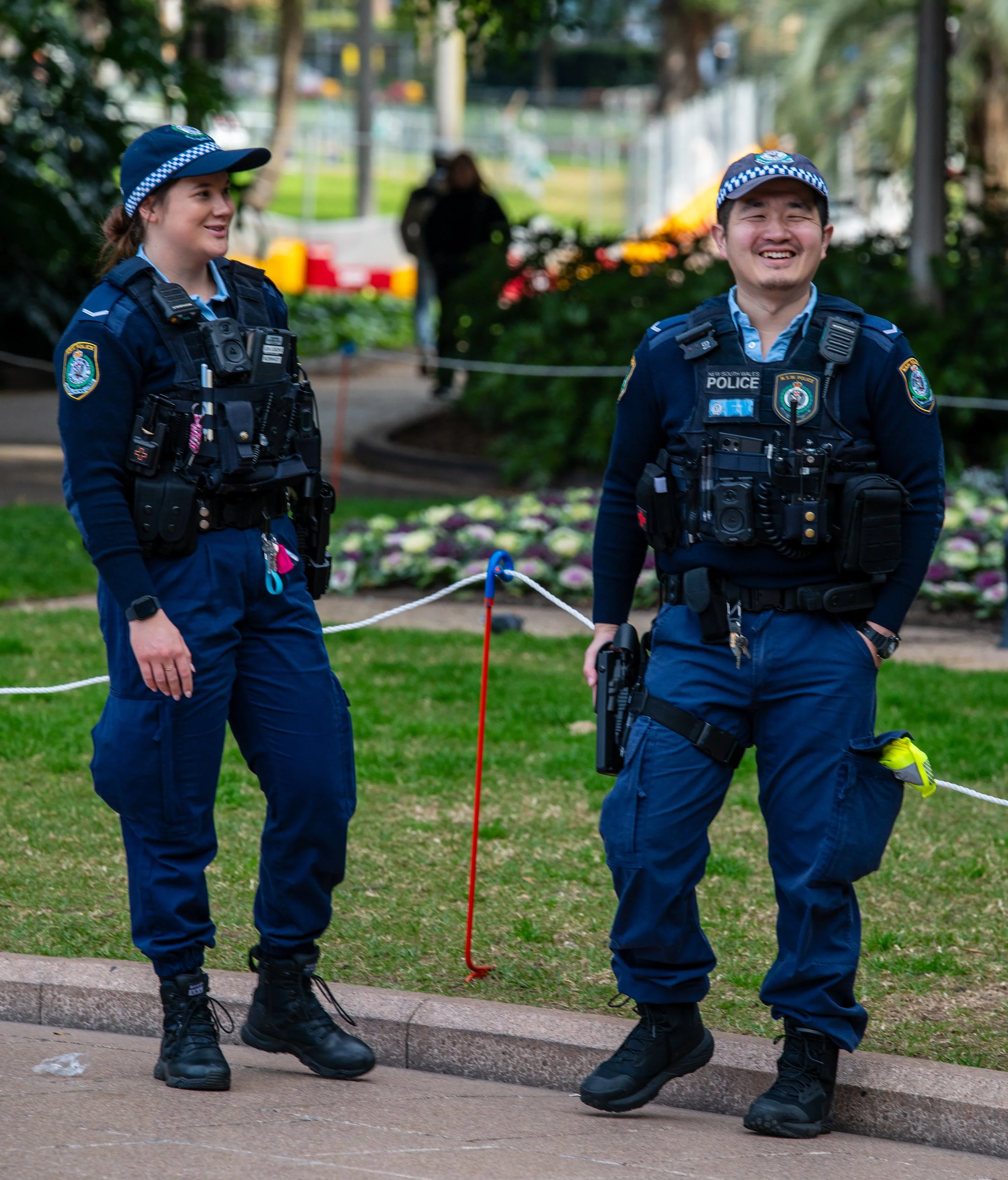 Two police officers, one woman and one man, standing outdoors on a grassy area, smiling and engaging in conversation. They are wearing navy blue uniforms with police badges, utility vests with equipment, and police hats. The background includes trees, flowers, and a person walking further away.
