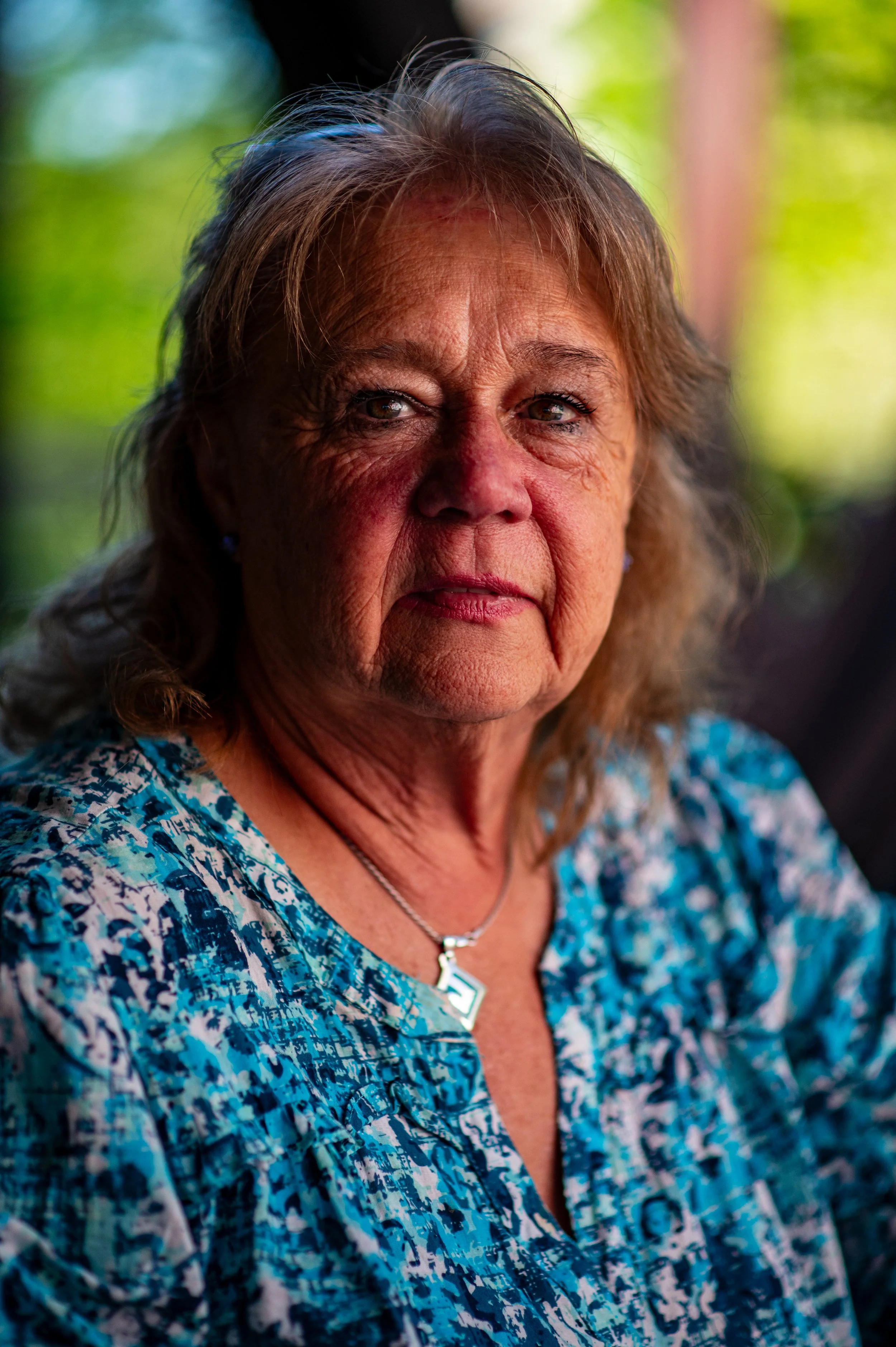 Close-up of an elderly woman with light brown hair and blue eyes, wearing a colorful blue and white patterned blouse, silver necklace, and earrings. She is outdoors with a blurred green background.