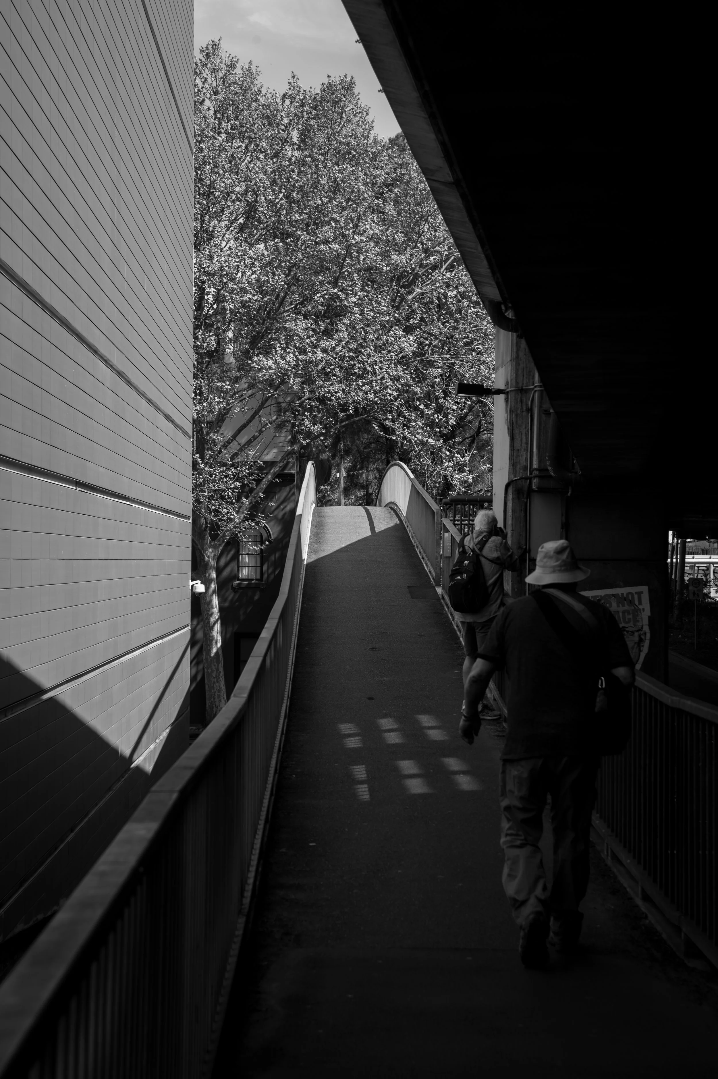 Two people walking along a narrow elevated walkway next to a building with trees in the background, sunlight casting shadows