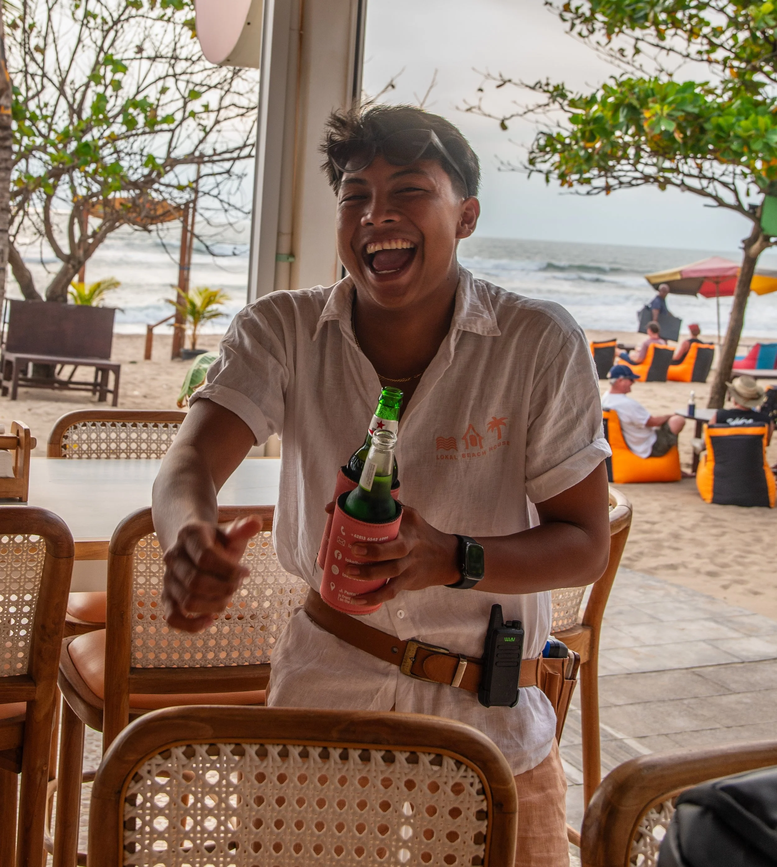 A young man with dark hair and sunglasses on his head, smiling and holding a beer bottle wrapped in a pink koozie, sitting at a table near the beach with trees, umbrellas, and people in the background.