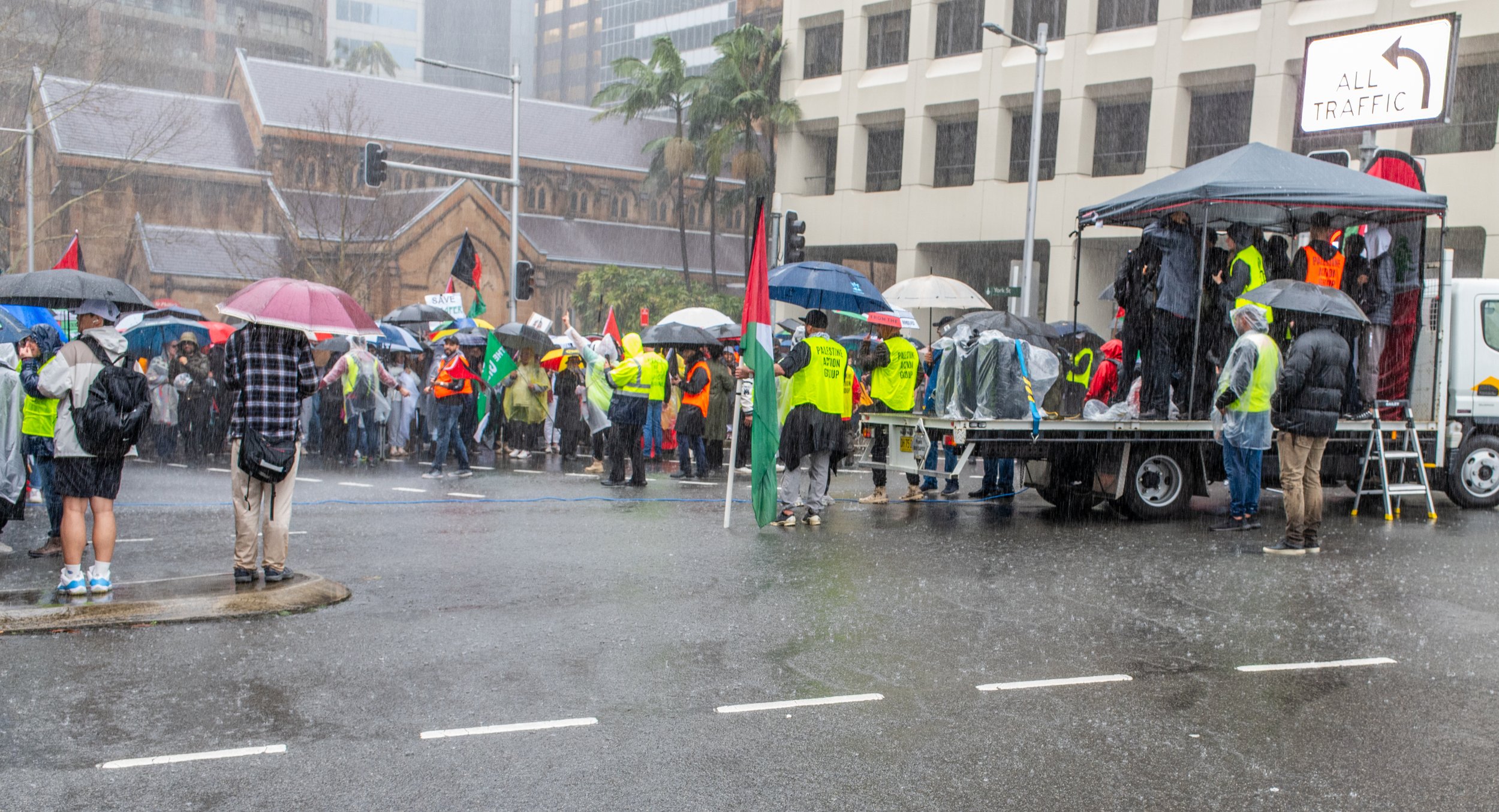 A crowd of protesters gathered on a rainy day, many holding umbrellas. Some individuals are wearing yellow vests with green text that reads "Palestinian Action Group." There is a truck with a canopy, where several people are speaking or organizing. Palestinian flags are visible among the protesters.