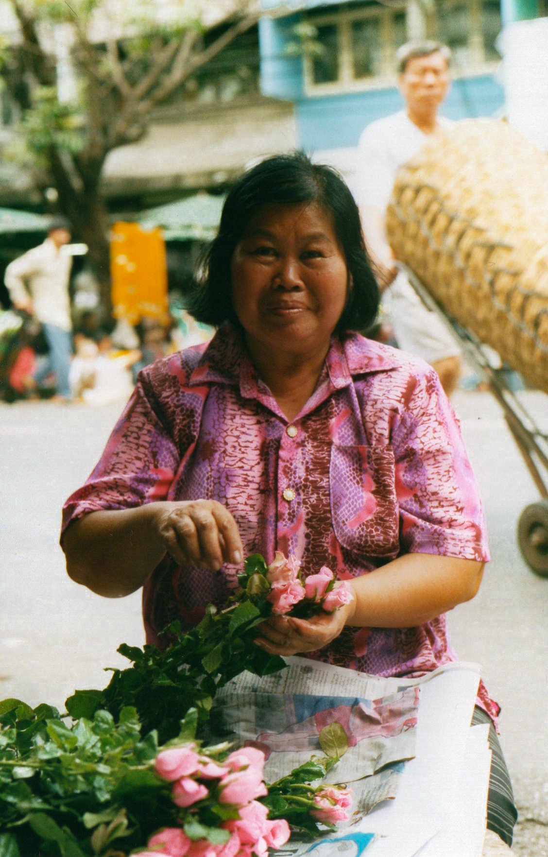 A woman in a pink patterned shirt holding pink flowers at an outdoor market with blurred people and a blue building in the background.