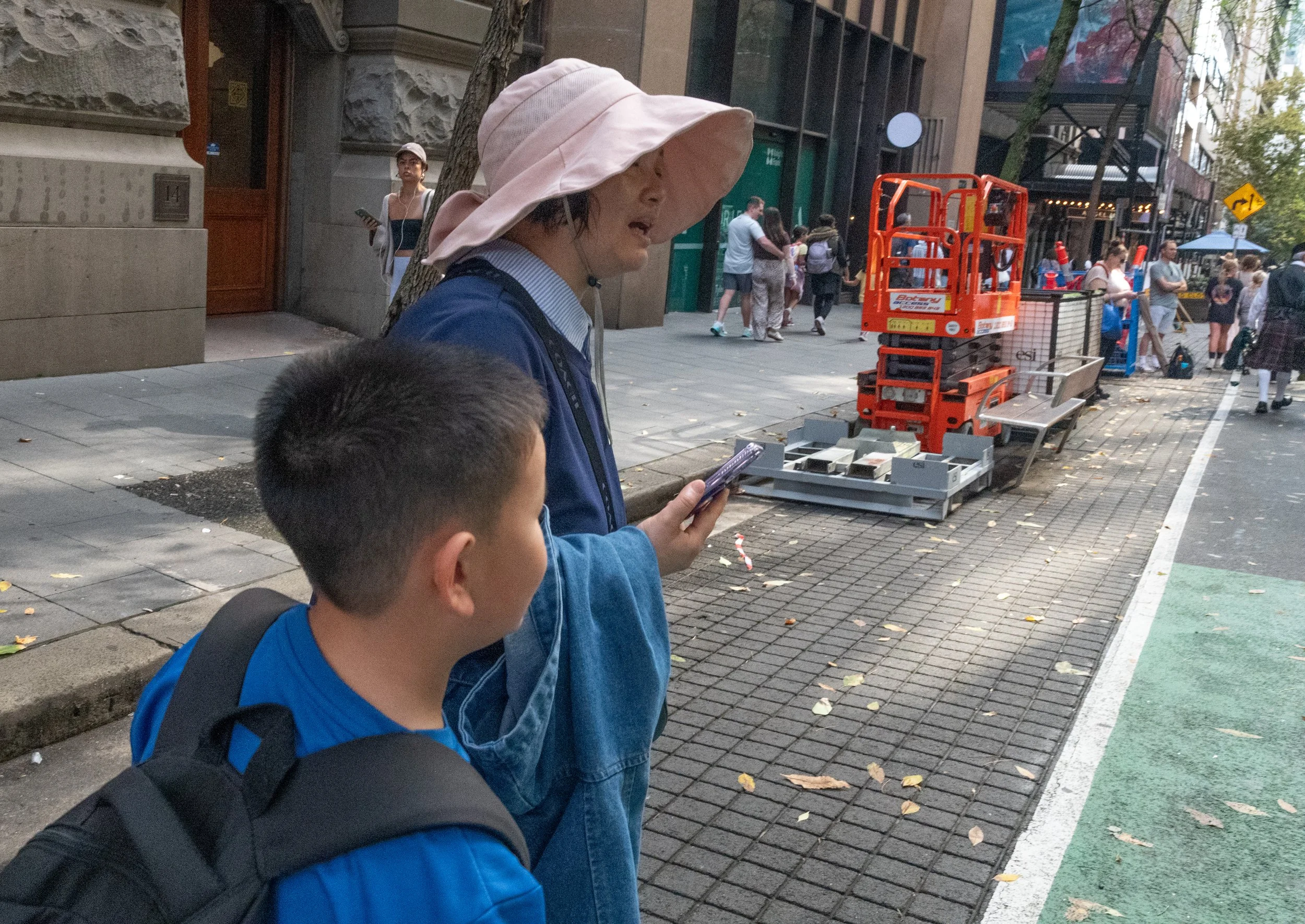 A woman wearing a pink wide-brimmed hat and a blue jacket stands on a city sidewalk, holding a smartphone, with a young boy in a blue shirt and backpack beside her. People walk in the background near construction equipment and a chair on a busy city street.