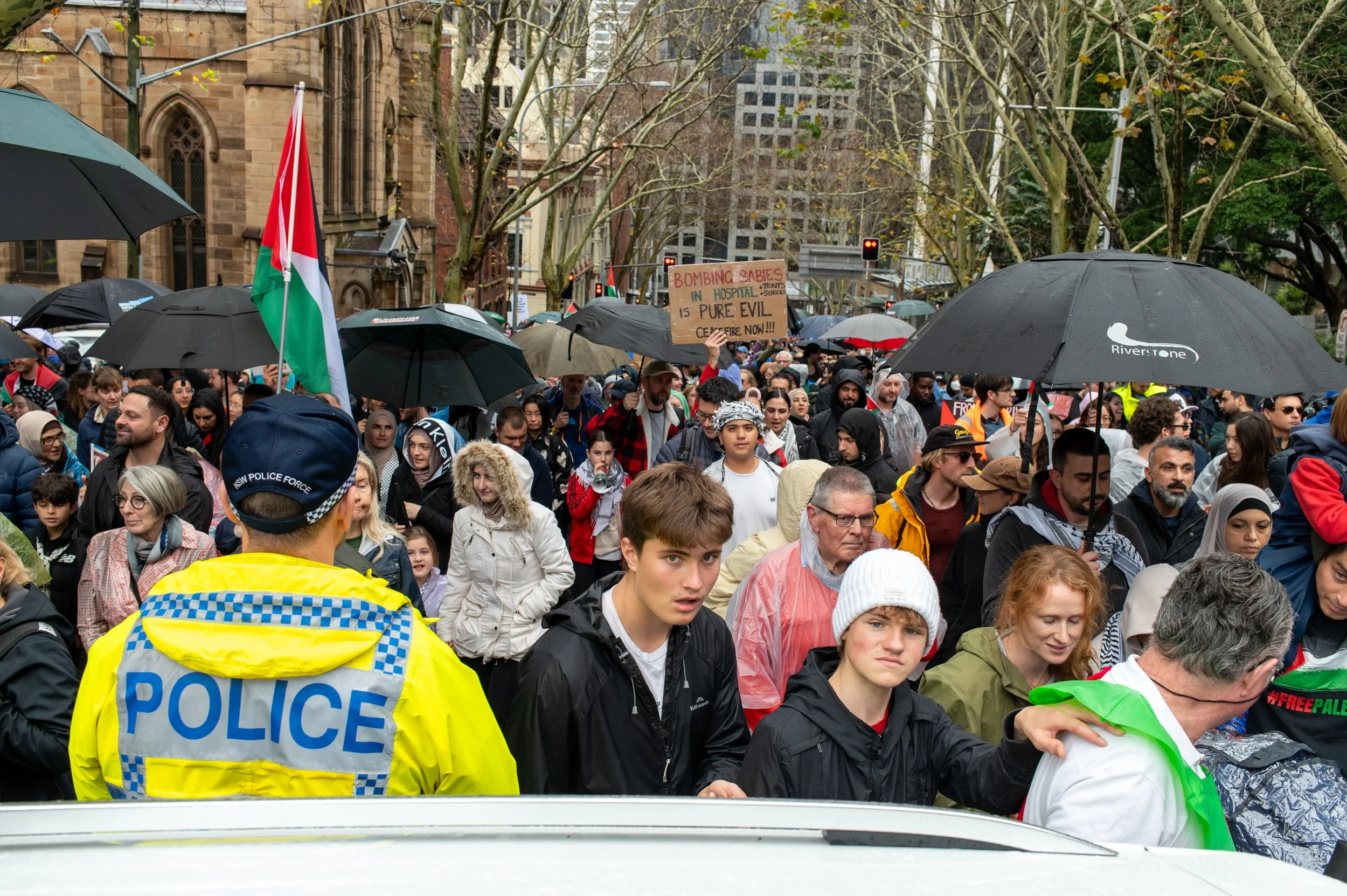Crowd of protesters holding umbrellas during a demonstration on a city street, with police presence and a protest sign about babies in hospital.