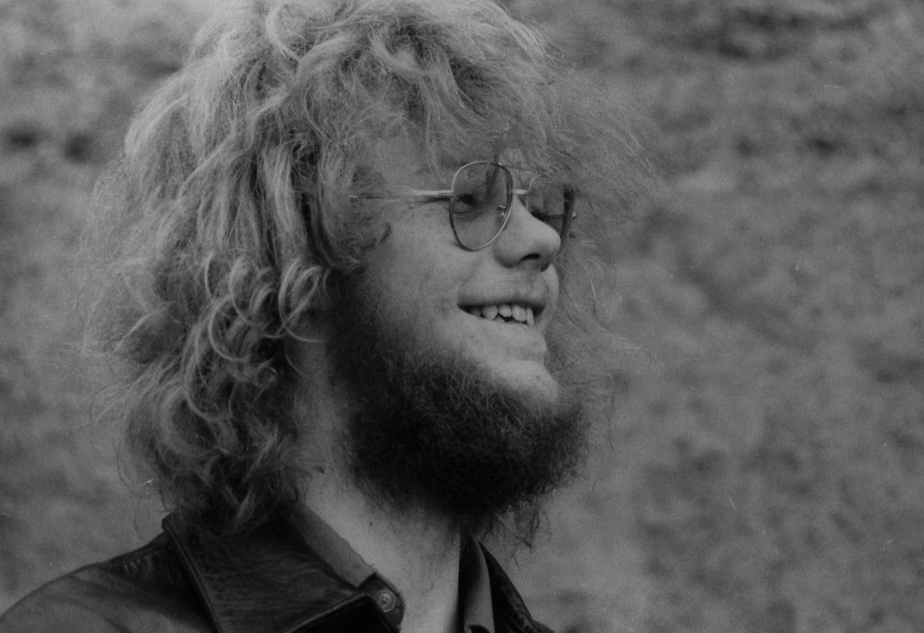 Black and white photo of a man with curly hair, glasses, and a beard smiling outdoors.