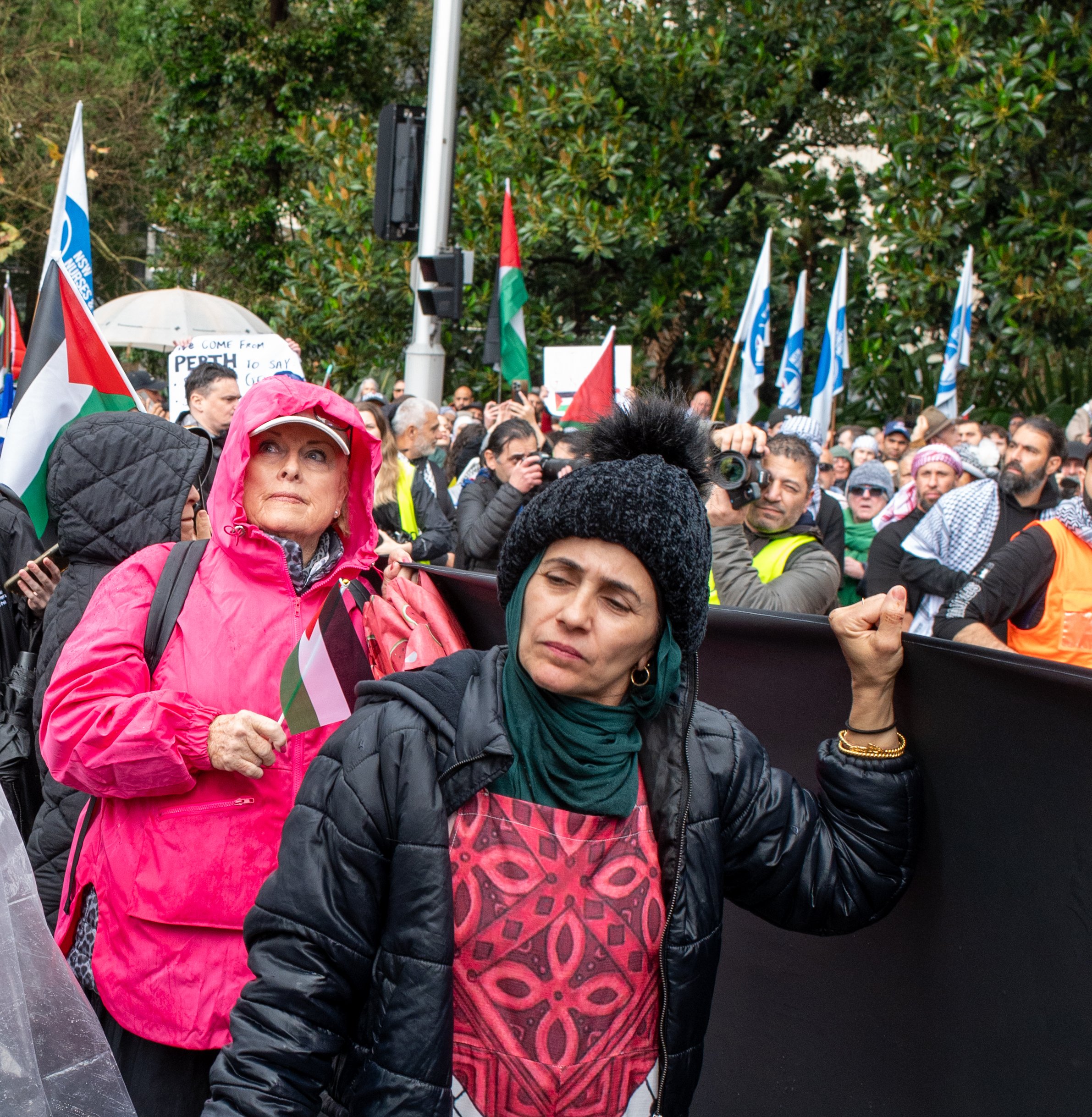 A group of people participating in a protest or rally, many holding flags and wearing warm clothing, with one woman in the foreground wearing a black jacket and green scarf, and another woman in a pink jacket with a hood, holding a small flag.