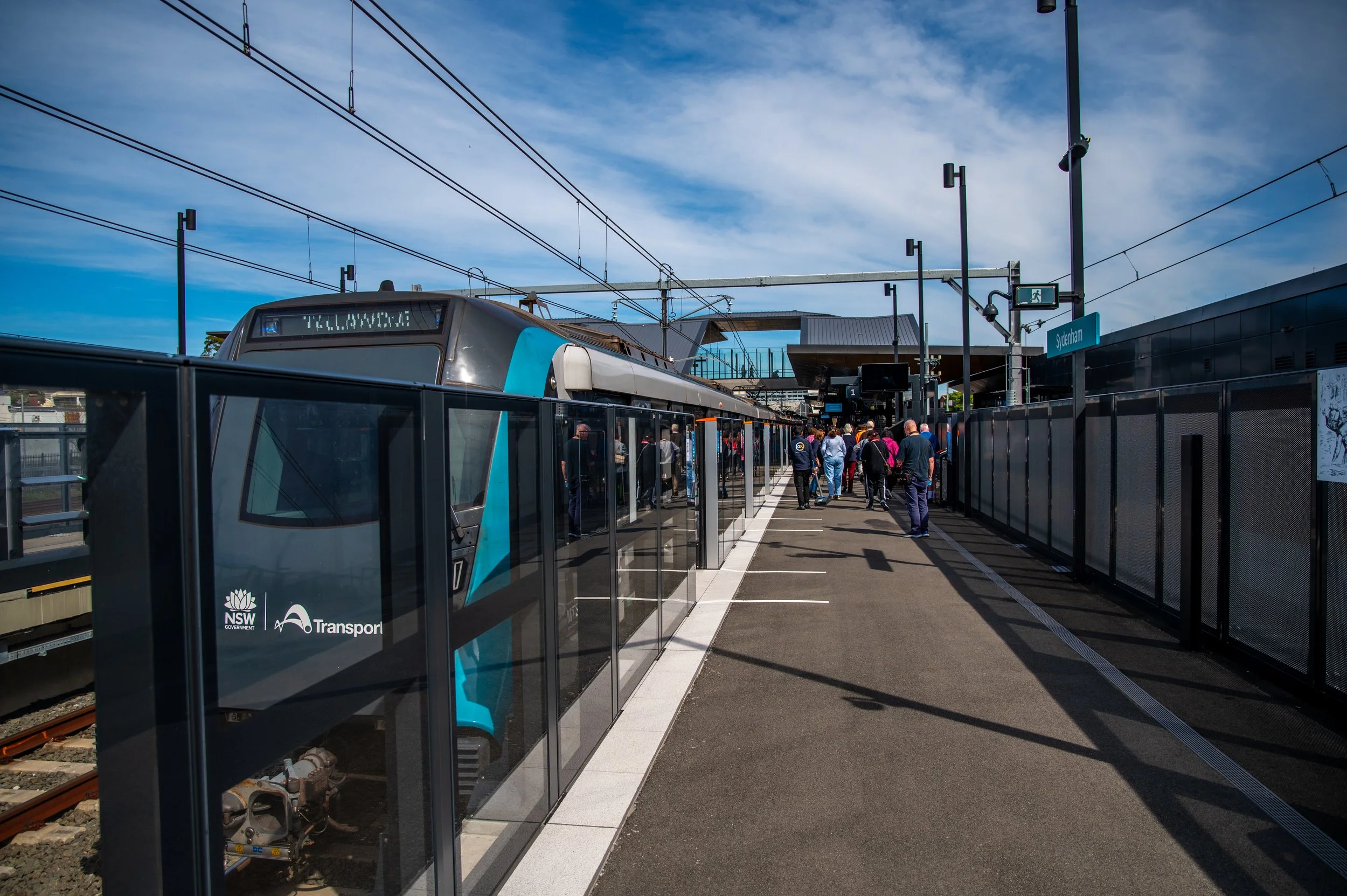 A train at a station platform with people waiting and walking, overhead electrical wires, and a sign indicating the station name as Sydenham.