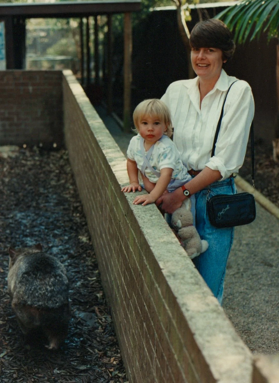 A woman and a young child leaning on a brick wall, observing a mole on the ground, outdoors.