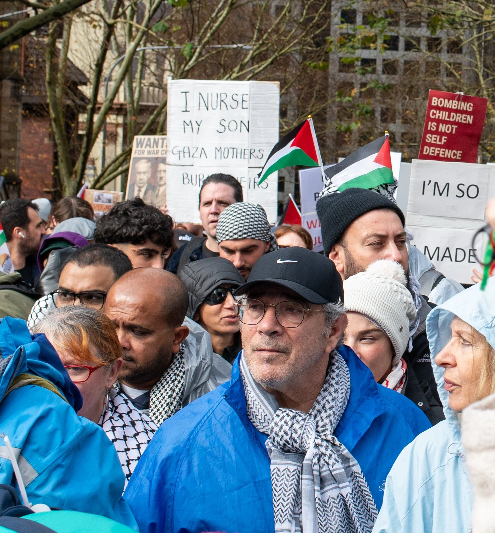 A crowd of people at a protest or rally, holding signs in support of Palestine, with some Palestinian flags visible.