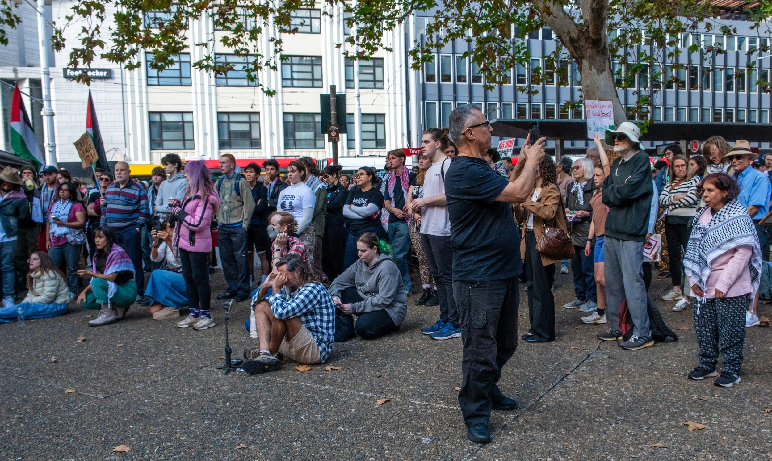 A diverse group of people gathered outdoors for a protest or rally, some standing, some sitting, with flags, signs, and a man in black taking a photo or video with his phone.