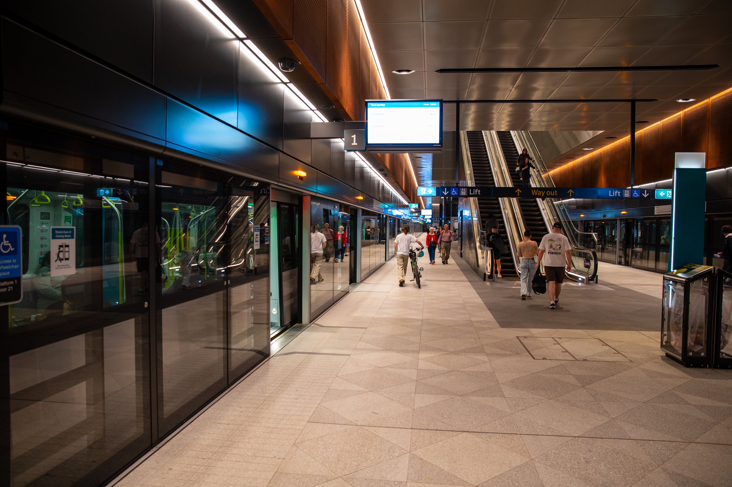 Indoor train station with escalators connecting to another level, people walking, some carrying luggage, and glass doors reflecting interior lights.