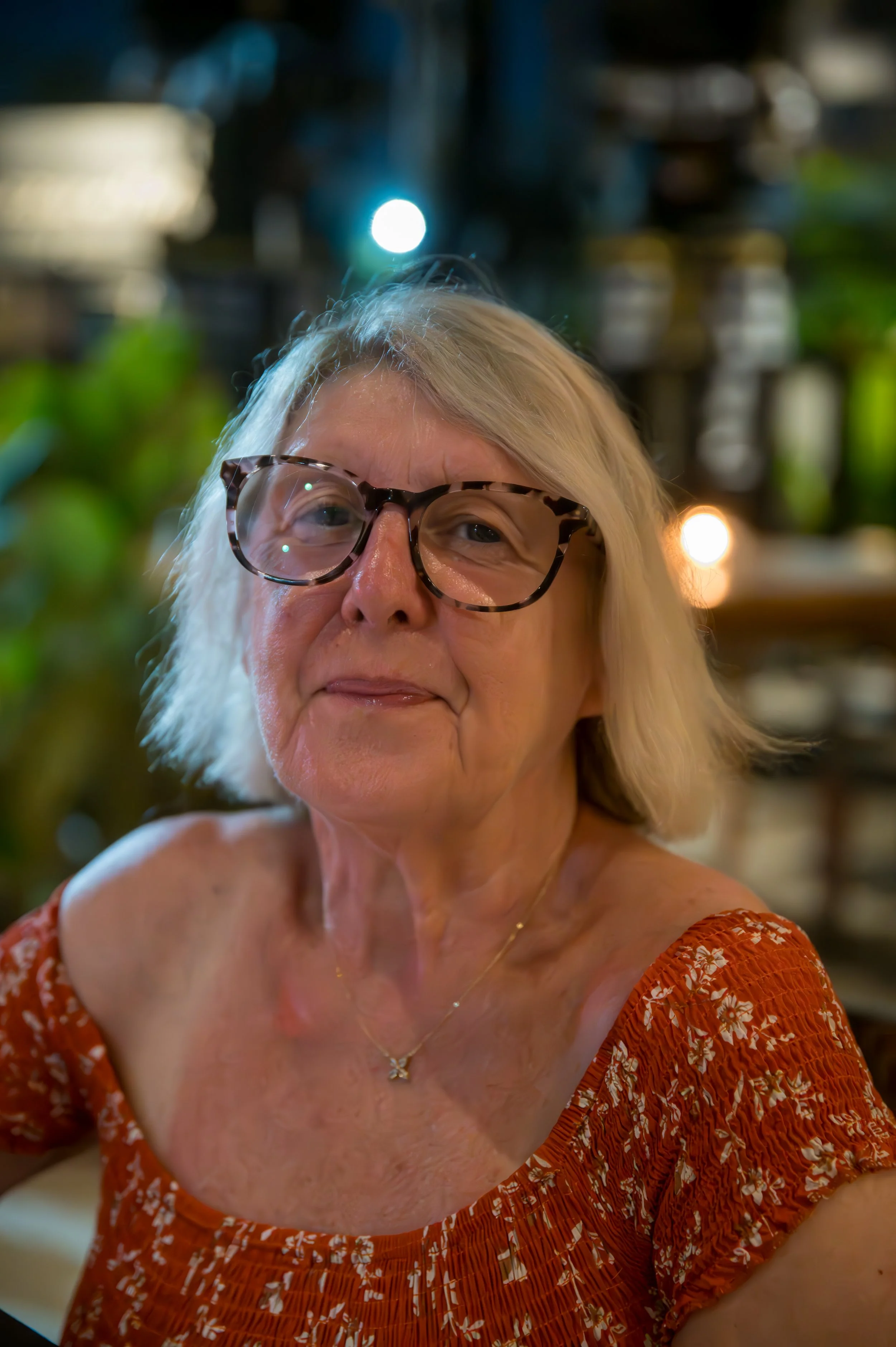 Close-up of a woman with blonde hair wearing glasses and a floral orange dress, smiling indoors with blurred background and bokeh lights.