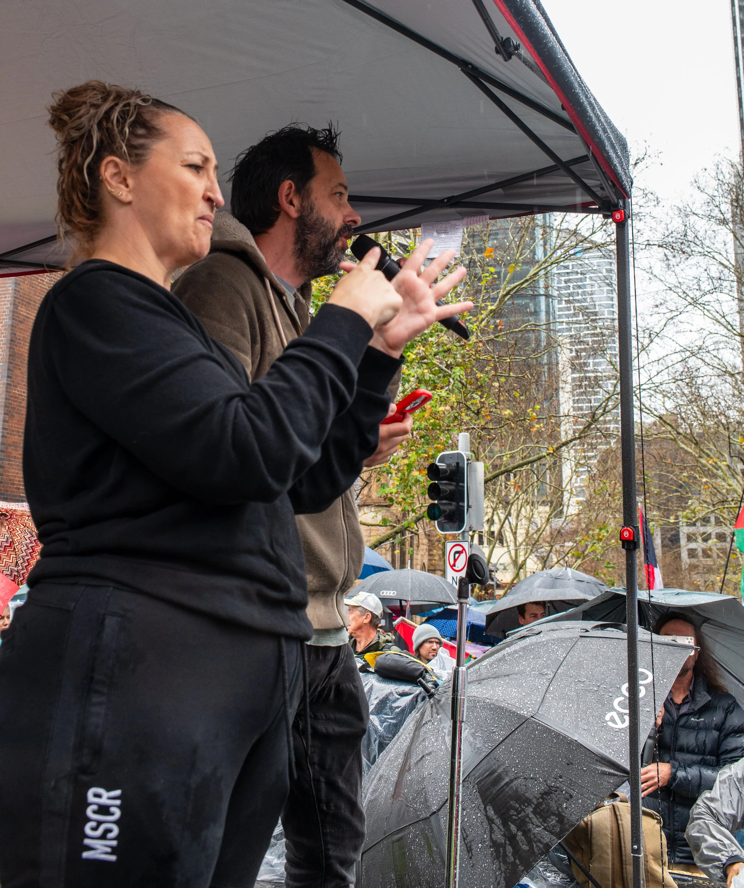 A woman and a man speaking at an outdoor event under a canopy, with a crowd holding umbrellas in the background in rainy weather.