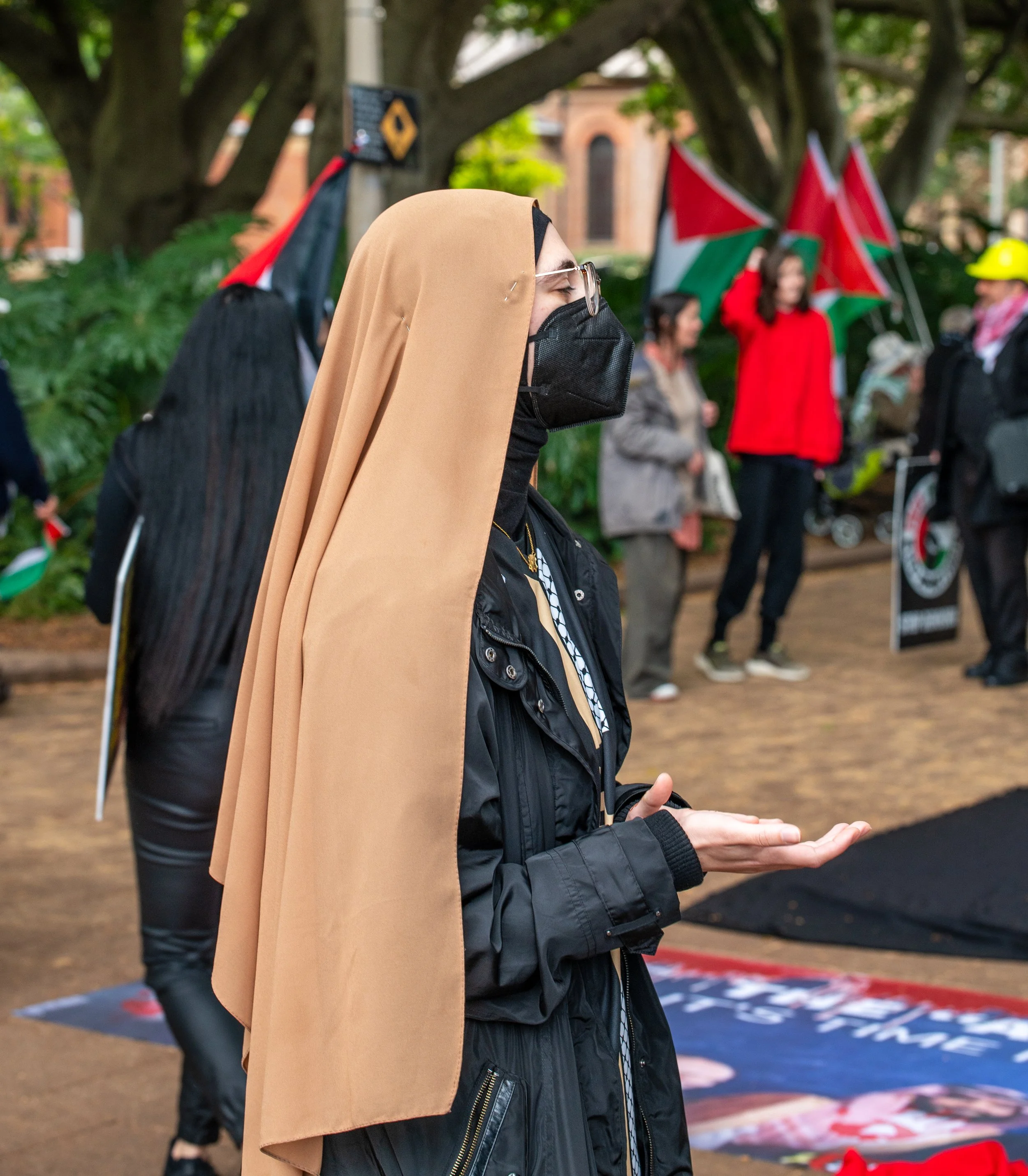 A woman wearing a black face mask and glasses stands outdoors during a gathering. She is dressed in a black jacket and is praying with her hands cupped. In the background, people hold Palestinian flags, and some are wearing masks and hats.