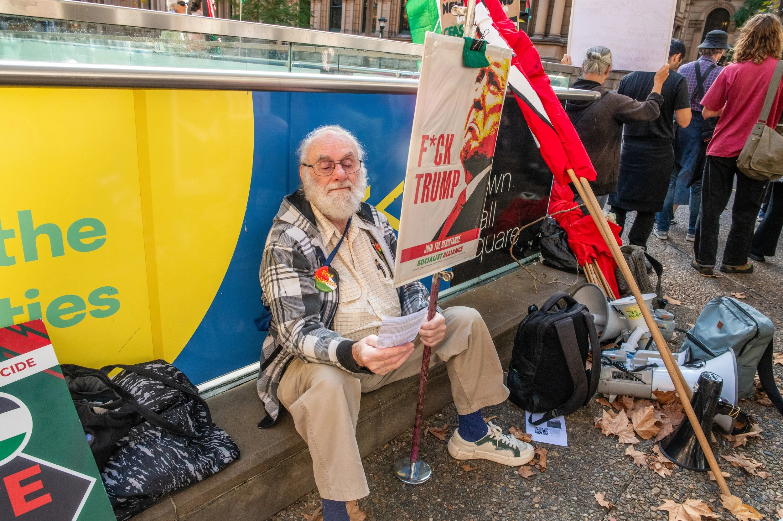 An older man with white hair and a beard, wearing glasses, a plaid jacket, and beige pants, is sitting on the ground holding a sign that reads 'F*CK TRUMP' at a political protest. Various signs, backpacks, a megaphone, and protest equipment surround him. There are other protesters standing in the background.
