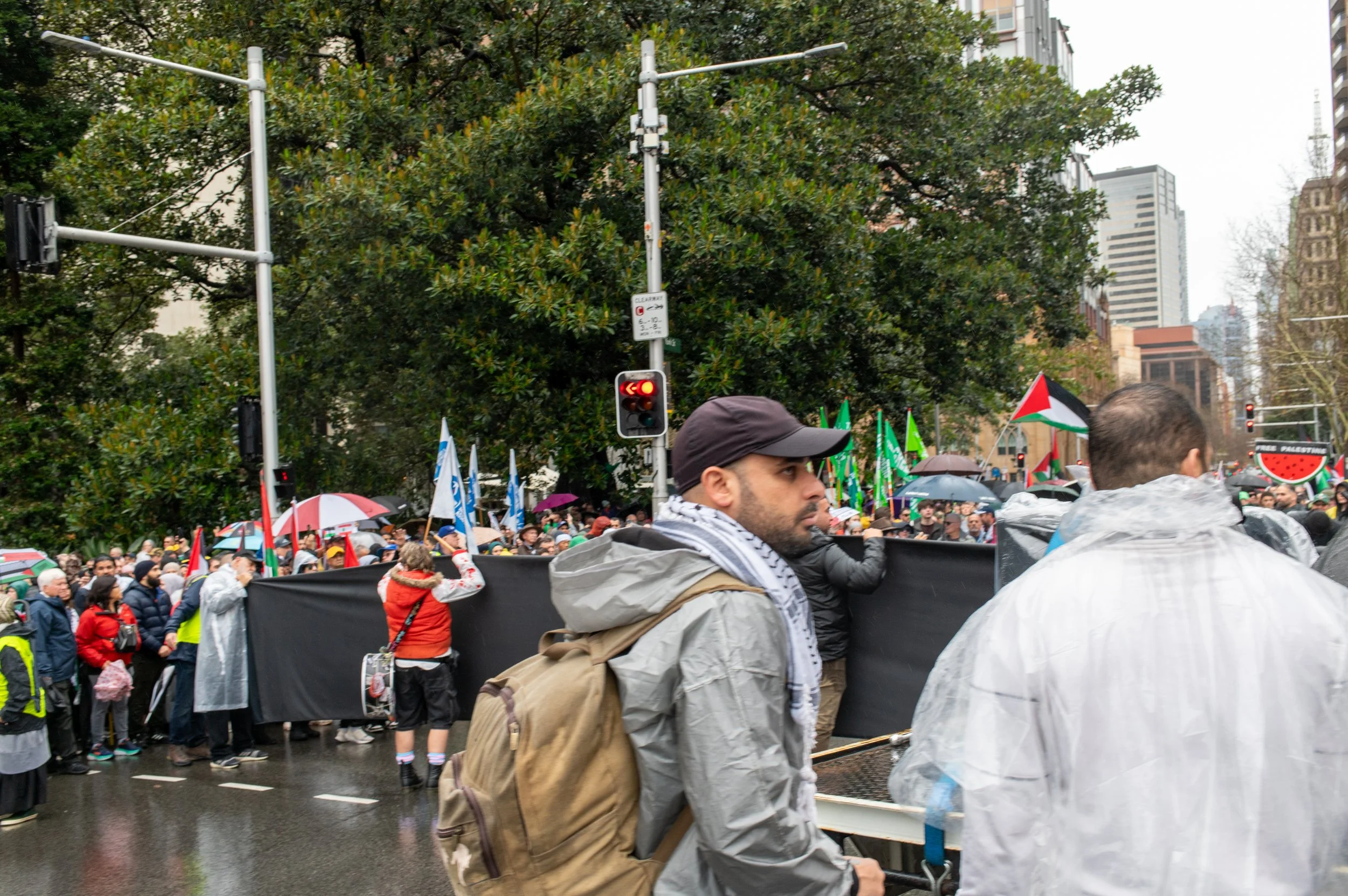 A large crowd of protesters gathered on a city street holding flags and banners, with some wearing rain jackets and holding umbrellas, amidst tall buildings and trees.