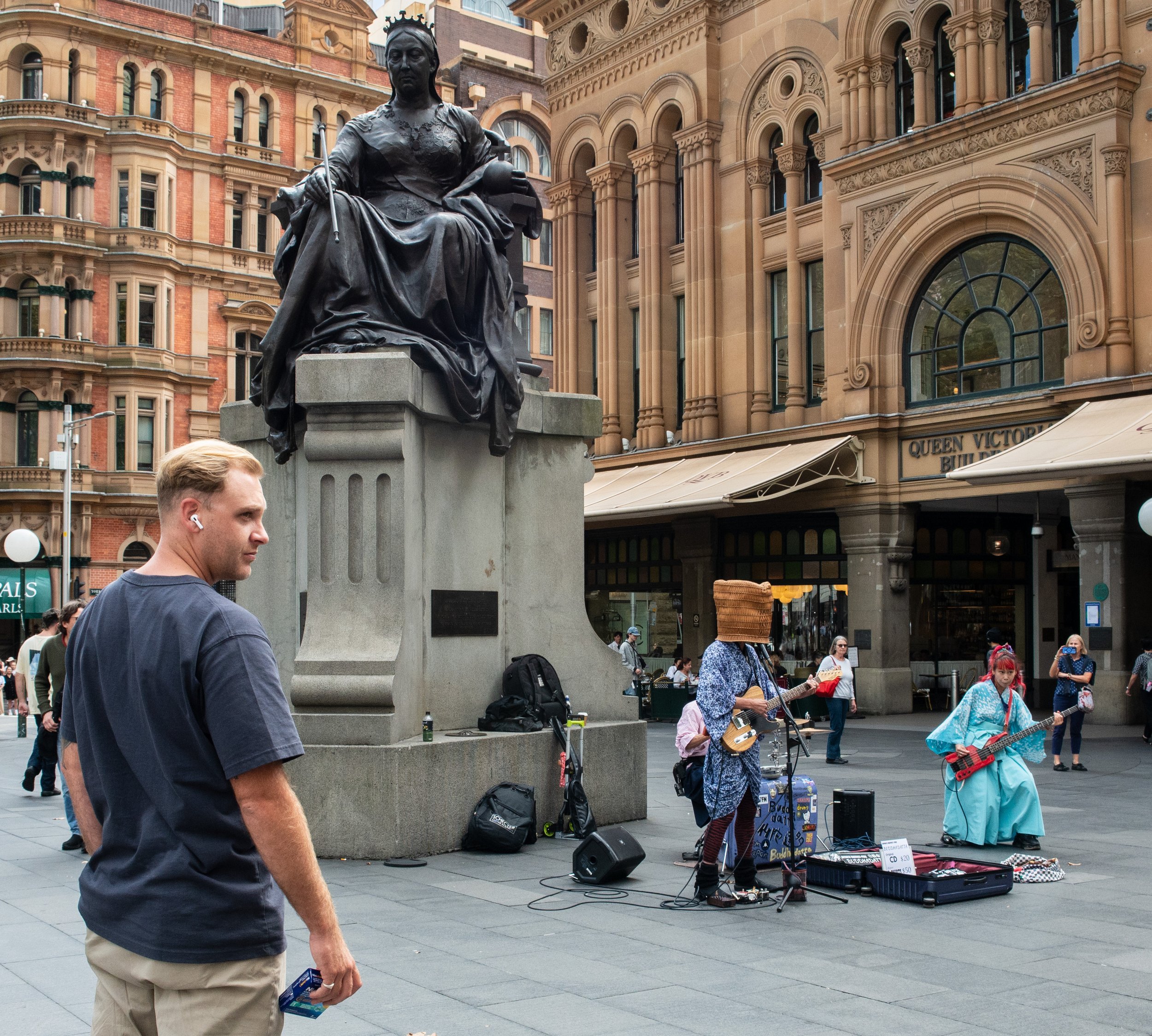A street scene featuring a large bronze statue of a seated woman on a stone pedestal, with a street musician and a female musician performing nearby. The background includes historic buildings and pedestrians.