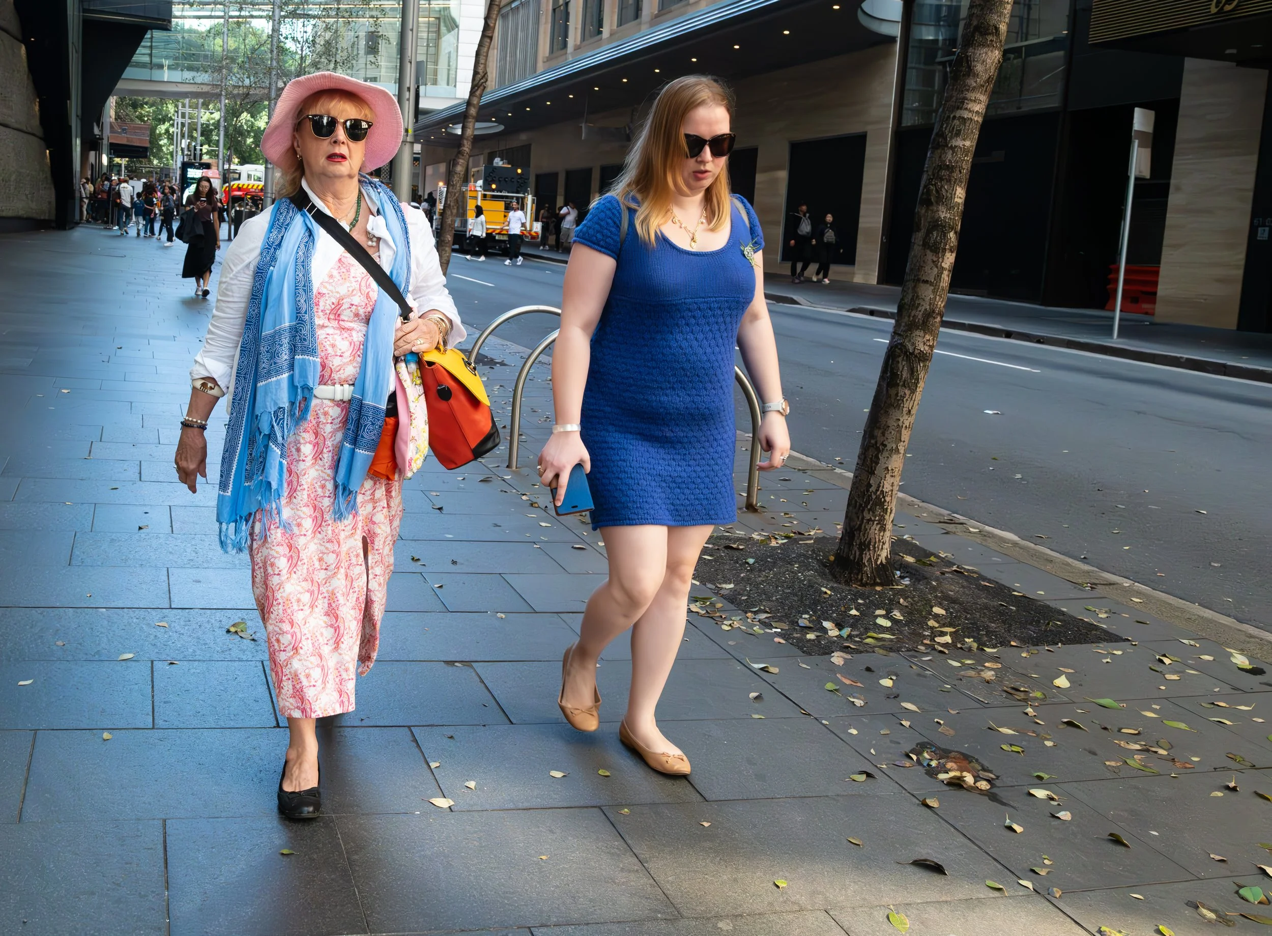 Two women walking on city sidewalk, one older woman in pink hat, sunglasses, and floral pants, and a younger woman in a blue dress and sunglasses, with trees and buildings in the background.
