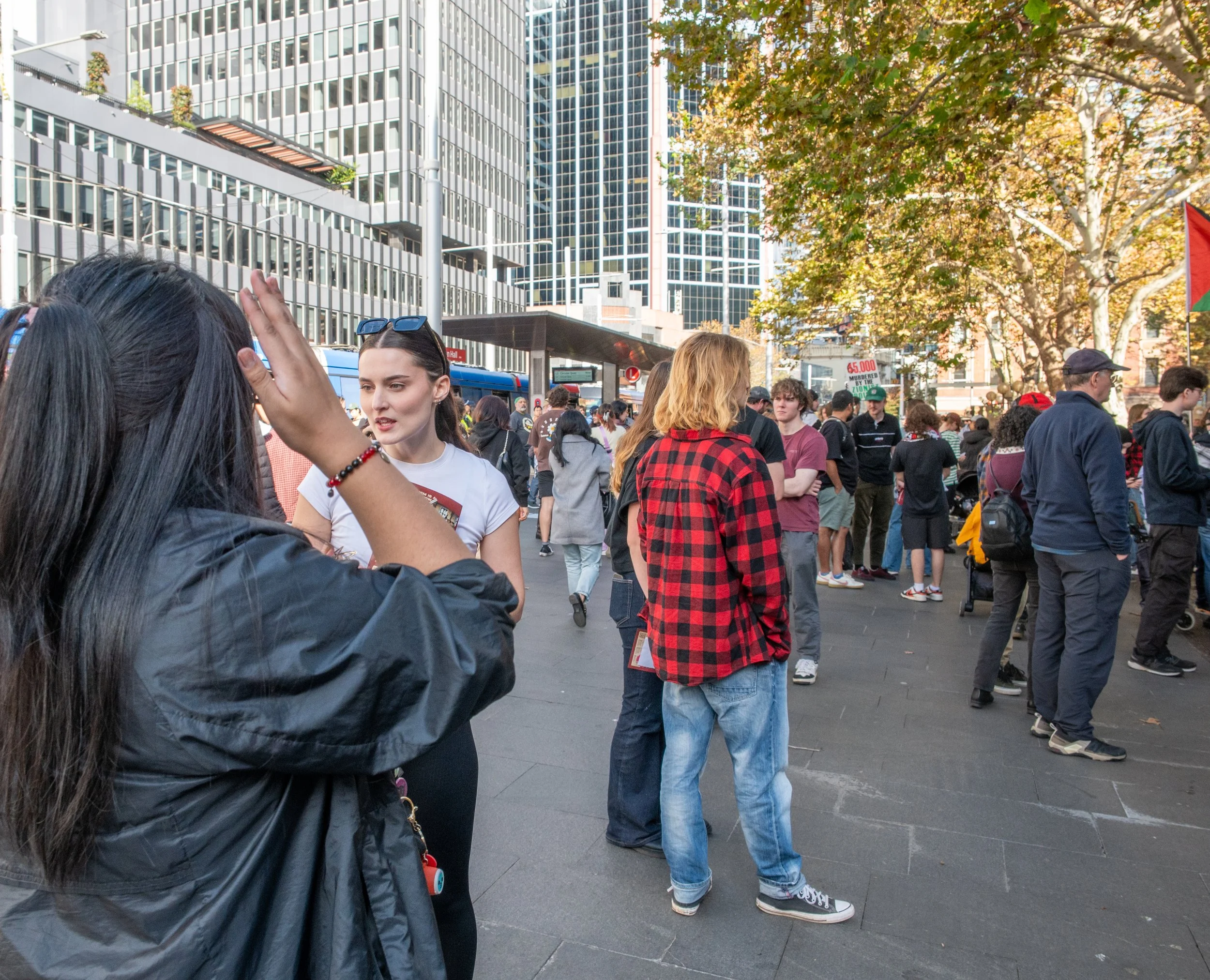 Crowd of people gathered outdoors in an urban setting with tall buildings, trees with fall foliage, and a bright sky.
