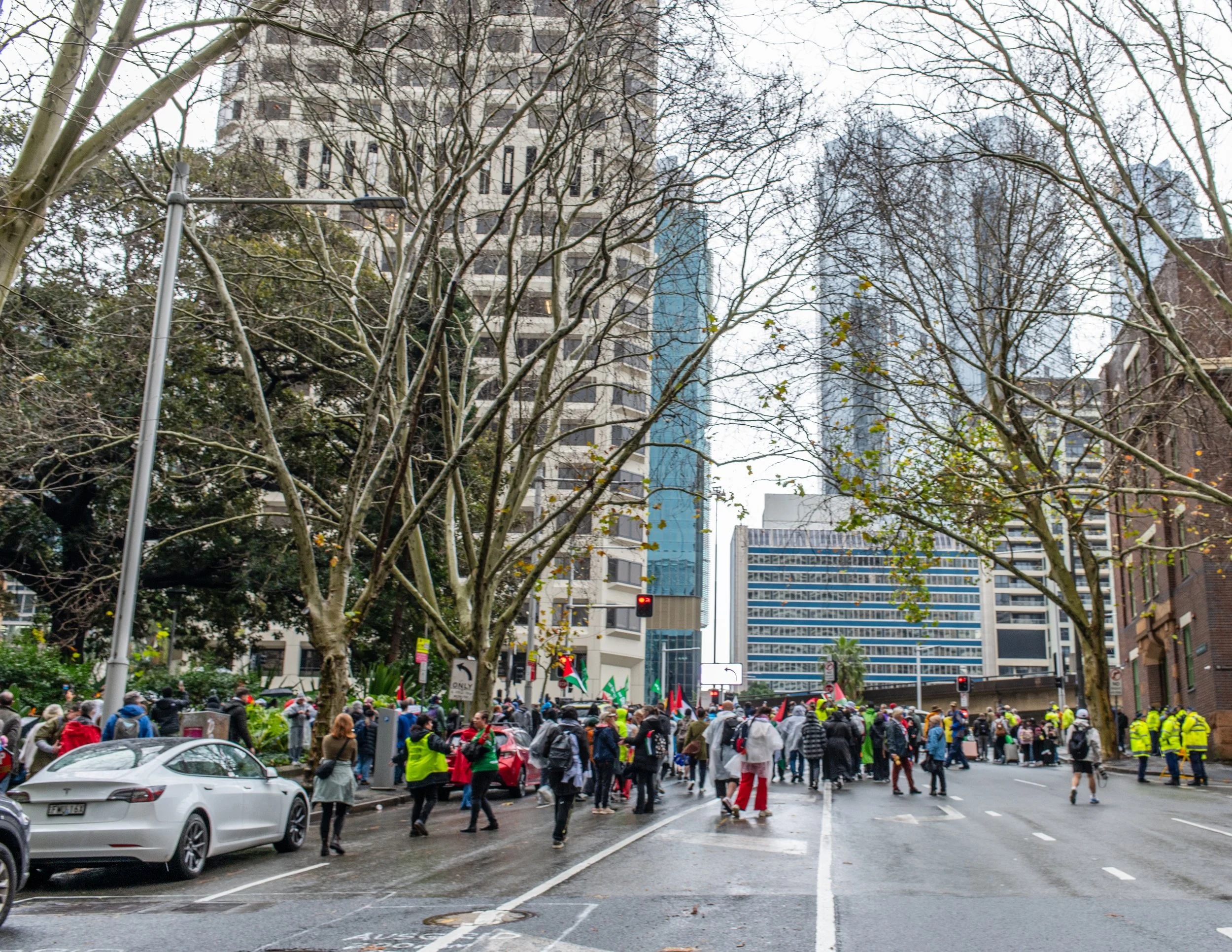 City street protests with people gathering, some wearing yellow vests, in an urban area with tall buildings and trees.