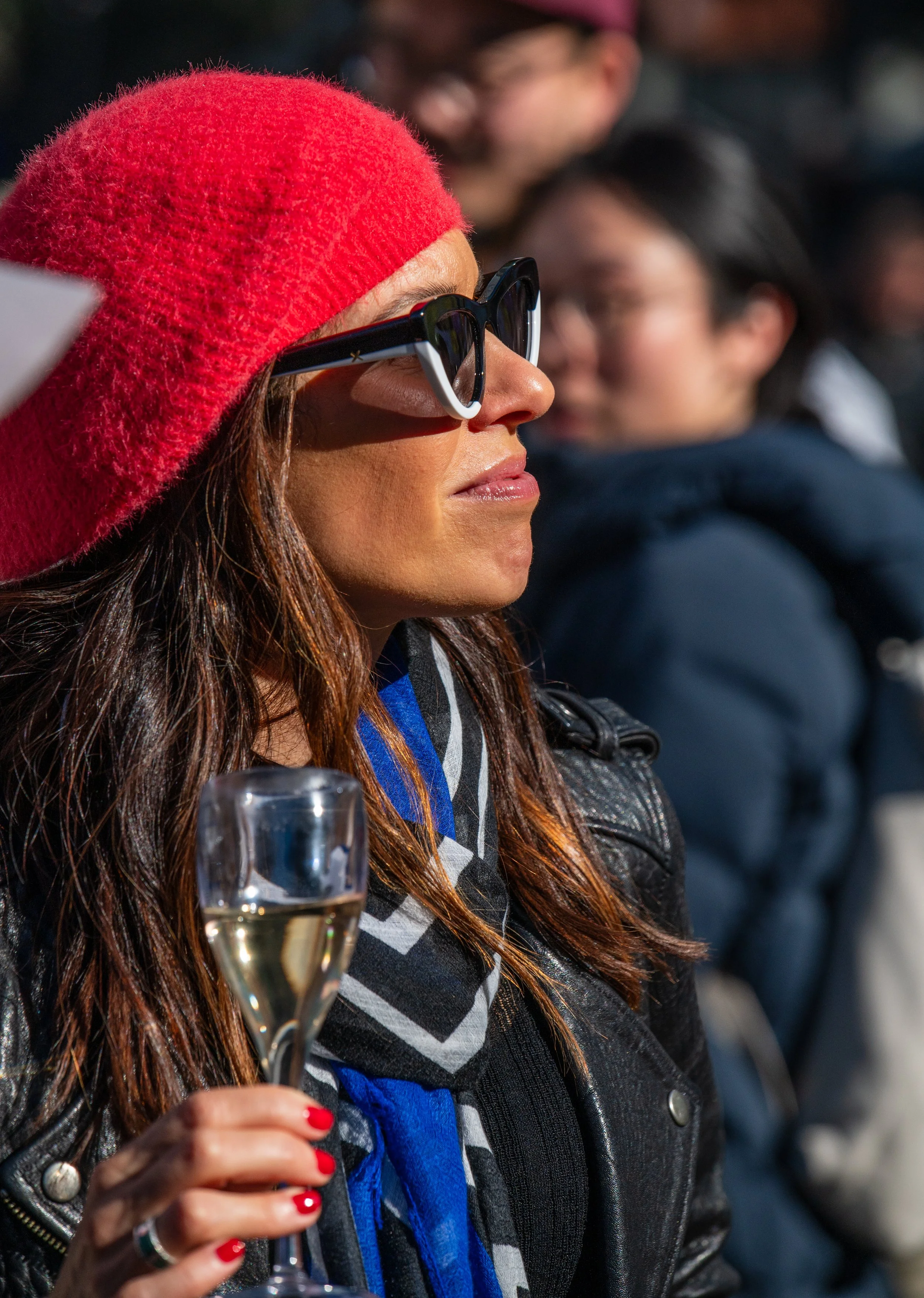 A woman with long brown hair, wearing a red knit hat, black sunglasses, a black leather jacket, and a black, white, and blue scarf, is holding a glass of champagne at an outdoor gathering.