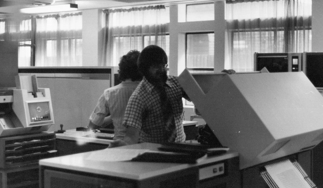 Two men working at desks in an office with large windows, using early computer equipment.