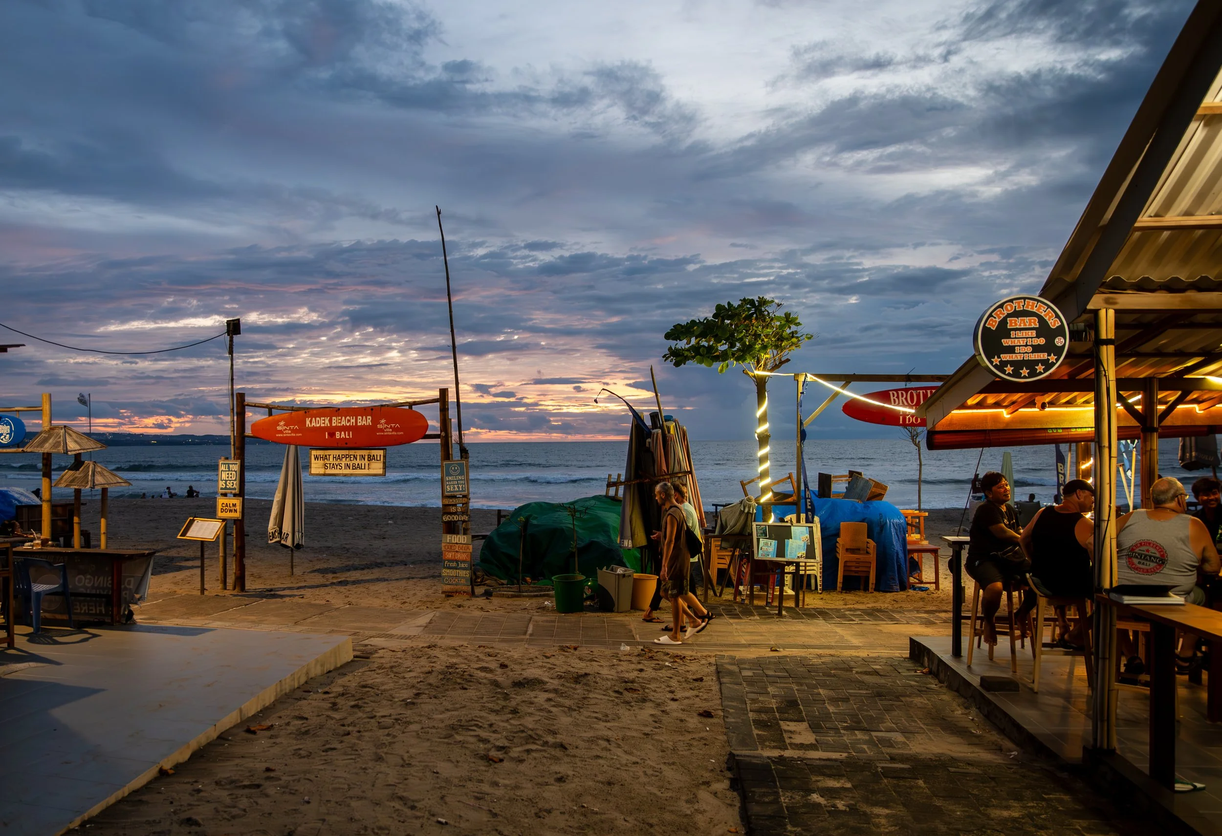 Beachside bar at sunset with tables, chairs, and people, with a view of the ocean and cloudy sky.