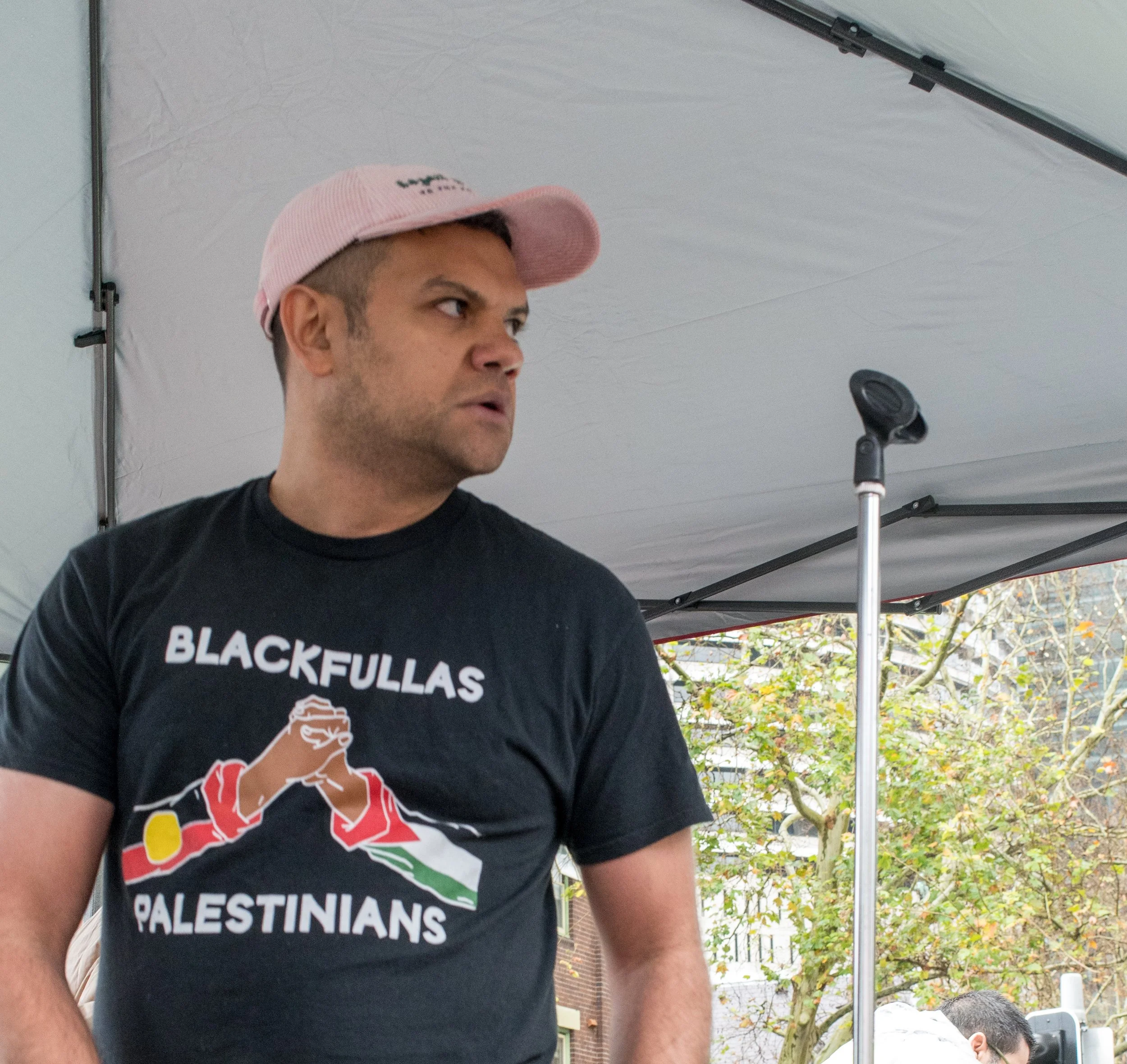 A man wearing a pink hat and a black T-shirt with the words 'Black Fullas Palestinians' and an illustration of hands holding a Palestinian flag, standing under a tent outdoors.